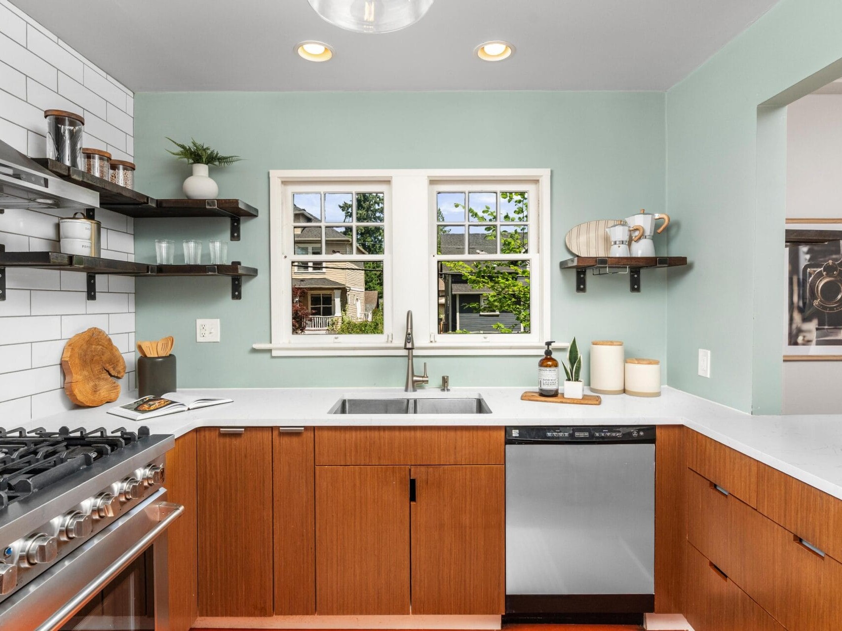 Modern kitchen with wood cabinets, white countertops, a stainless steel stove and dishwasher, open shelves with dishes, a large sink under two windows, and a mint green wall.