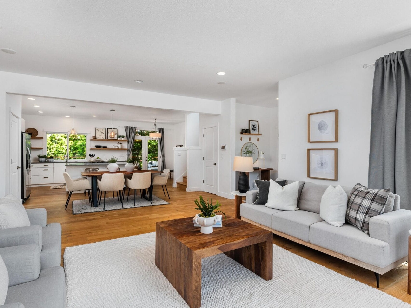 A modern open-concept living and dining area with light walls, wooden floors, a gray sofa, armchair, wooden coffee table, dining table with eight chairs, and a kitchen in the background. Large windows provide natural light.