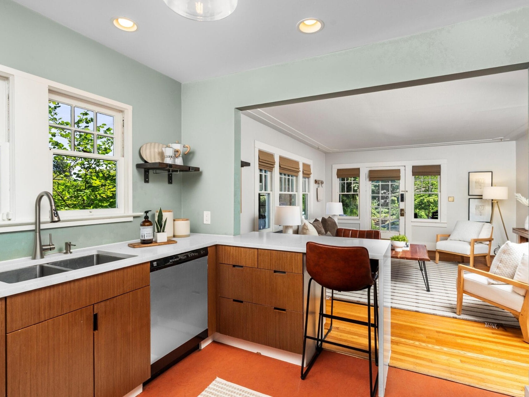 Modern kitchen with brown cabinets, white countertops, and a black dishwasher opens to a bright living room with large windows, a sofa, armchairs, and wood flooring. Green trees are visible through the kitchen windows.
