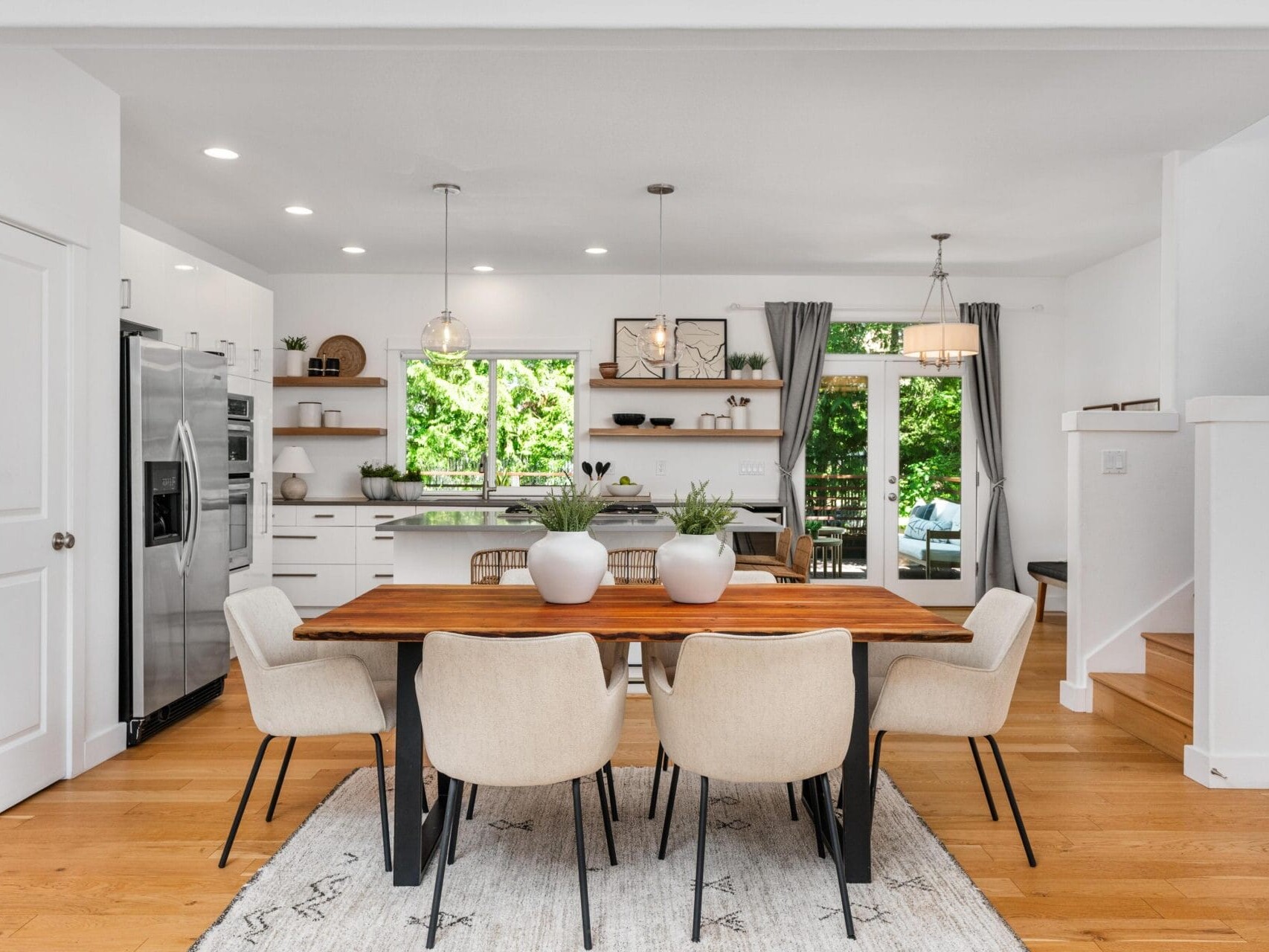 A modern open-concept kitchen and dining area with wooden floors, a wooden dining table, six beige chairs, potted plants, stainless steel appliances, white cabinets, and large windows letting in natural light.
