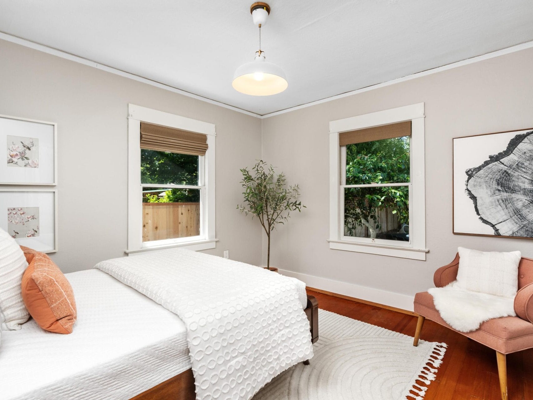 A bright bedroom with a bed, white bedding, orange pillows, a pink chair with a white rug, two windows, framed art on the walls, and a plant by the window. Natural light fills the room.