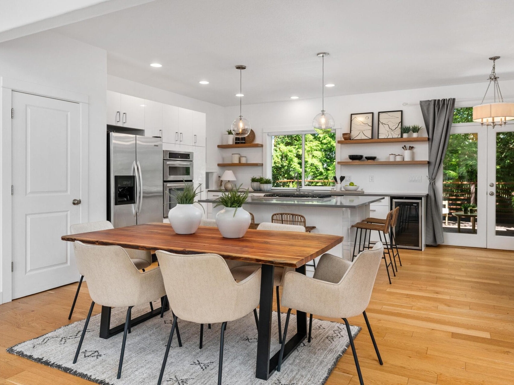 Modern open kitchen and dining area with a wooden table, beige chairs, a light rug, kitchen island with stools, stainless steel appliances, large windows, and glass doors opening to a green outdoor view.