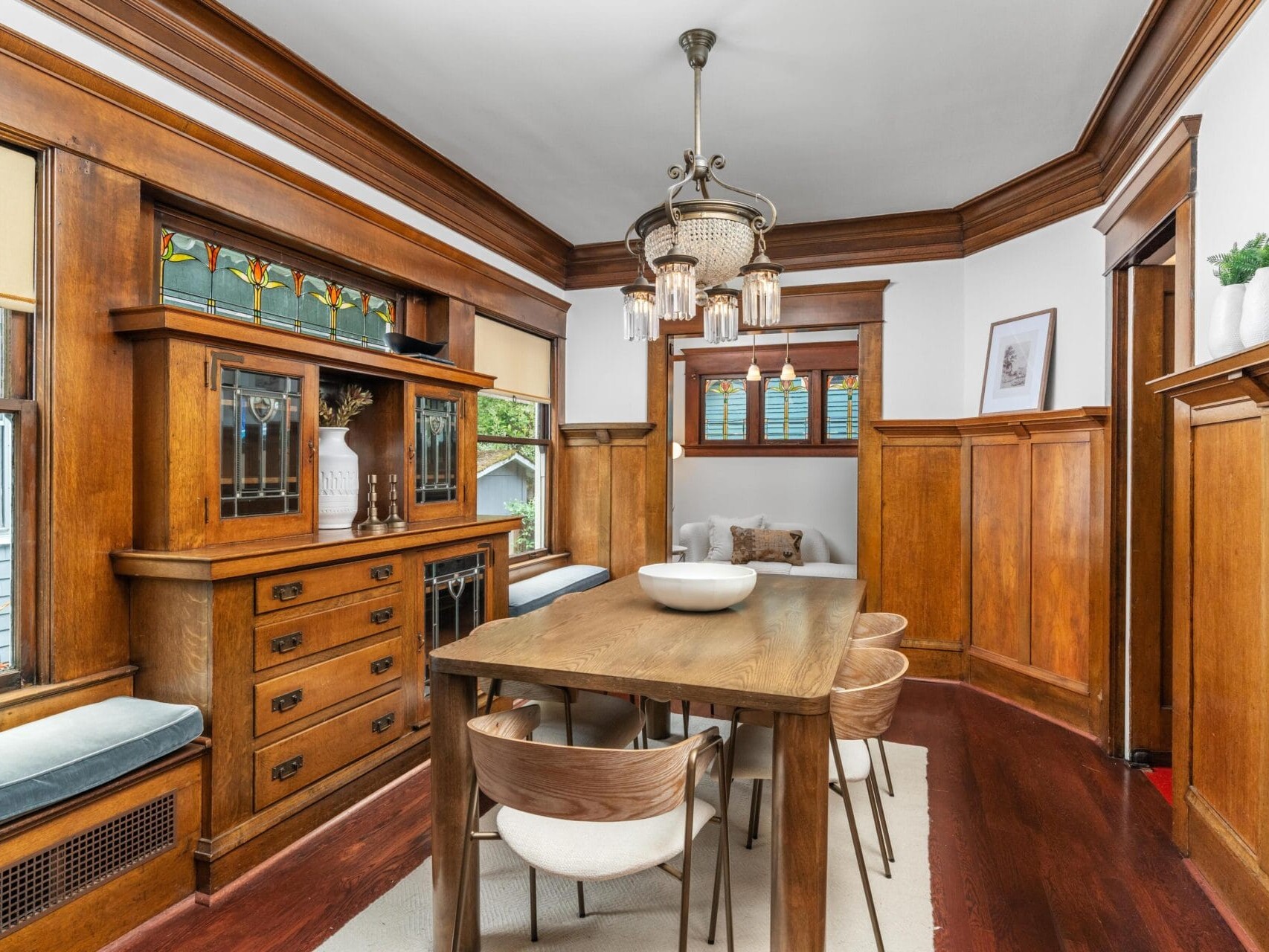 A dining room with warm wood paneling, a wooden table with six chairs, a built-in hutch with stained glass, cushioned window seats, and a vintage chandelier hanging from the ceiling.