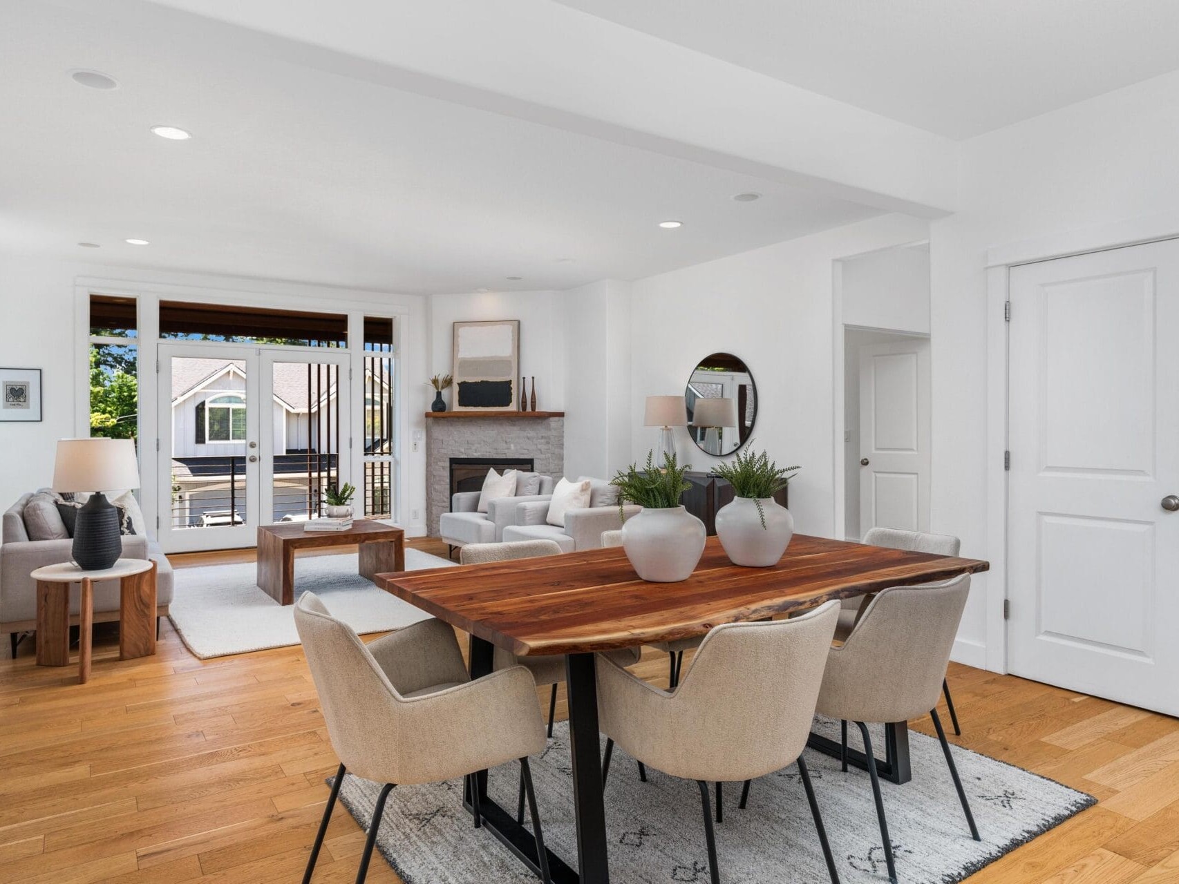 Open-concept living and dining room with light walls, wooden floors, a wooden dining table with six beige chairs, and a cozy seating area with sofas, a fireplace, large windows, and modern decor.