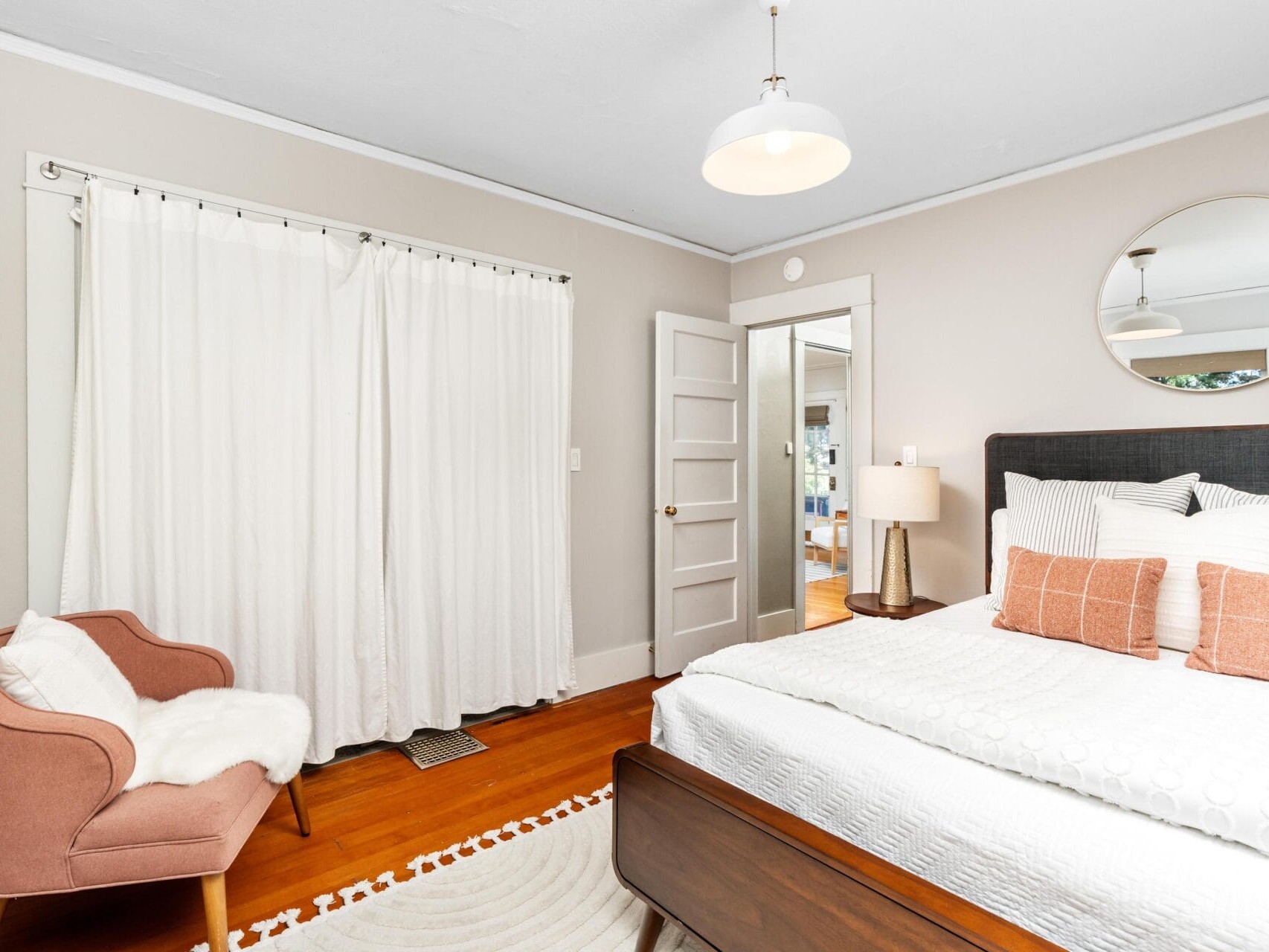 Bright bedroom with a modern design, featuring a bed with white and blush pillows, a round mirror, a pink armchair with a white throw, white curtains, hardwood floor, and a nightstand with a lamp.