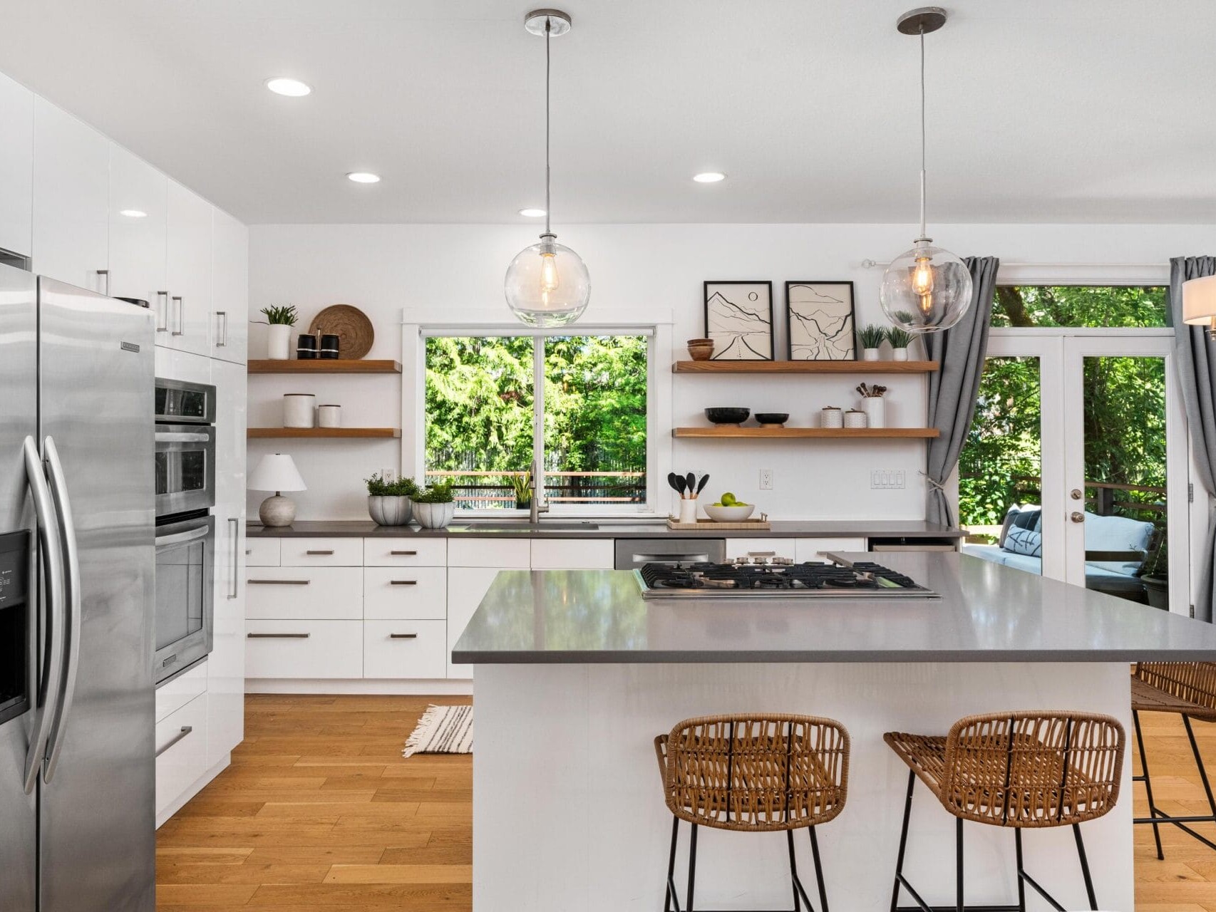 Modern kitchen with white cabinets, stainless steel appliances, wooden floors, open shelves, a large island with a gray countertop, wicker barstools, pendant lights, and large windows letting in natural light.