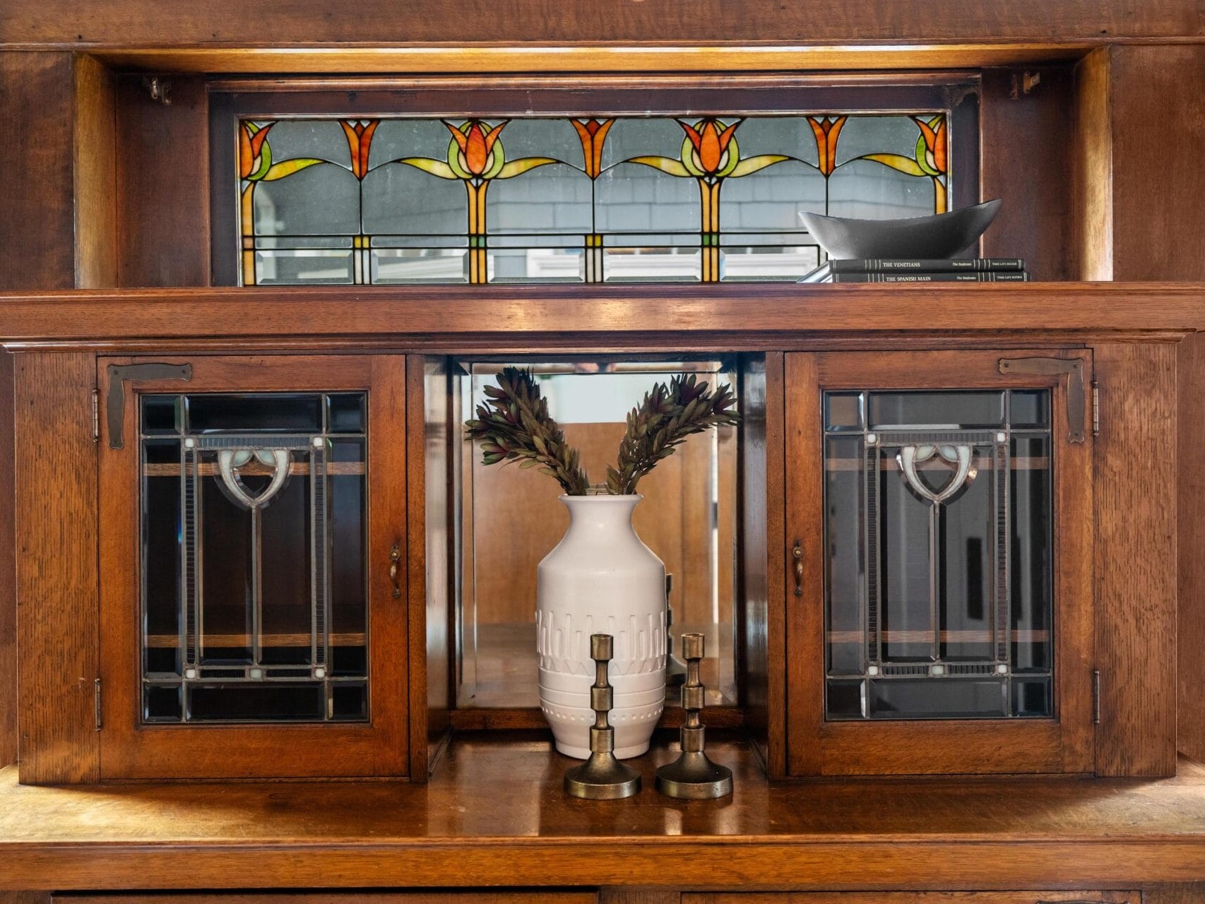 A wooden cabinet with stained glass doors and a colorful floral stained glass panel above, featuring a white vase with dried leaves and two candlesticks displayed in the center.