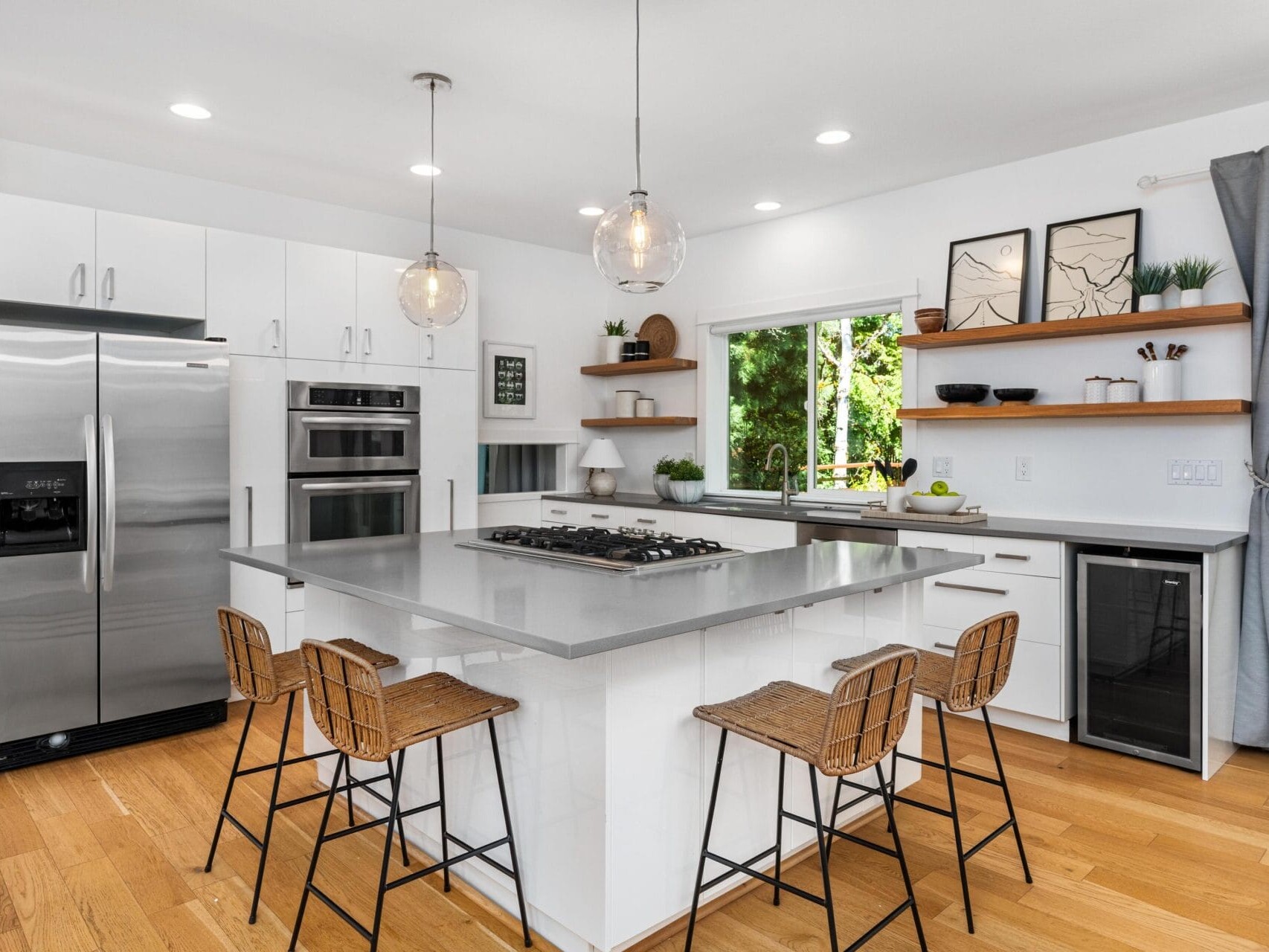 Modern kitchen with white cabinets, stainless steel appliances, an island with a built-in cooktop and four wicker barstools, open wooden shelves, pendant lights, and large window with gray curtain.