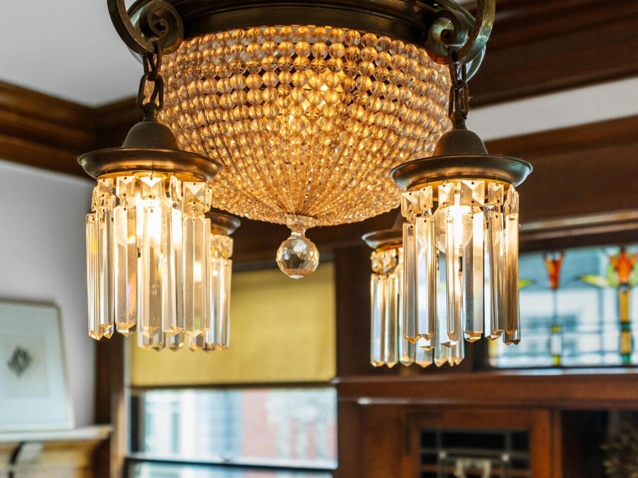 Antique bronze and crystal chandelier with four lights and hanging glass prisms, suspended from a ceiling in a room with wood trim, a window, and stained glass details in the background.