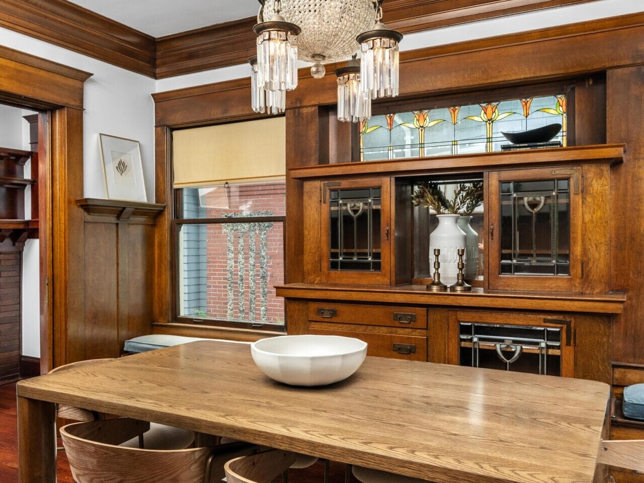 A dining room with a wooden table, four modern chairs, a white bowl centerpiece, a vintage wooden hutch with stained glass, large windows, and a decorative chandelier overhead.