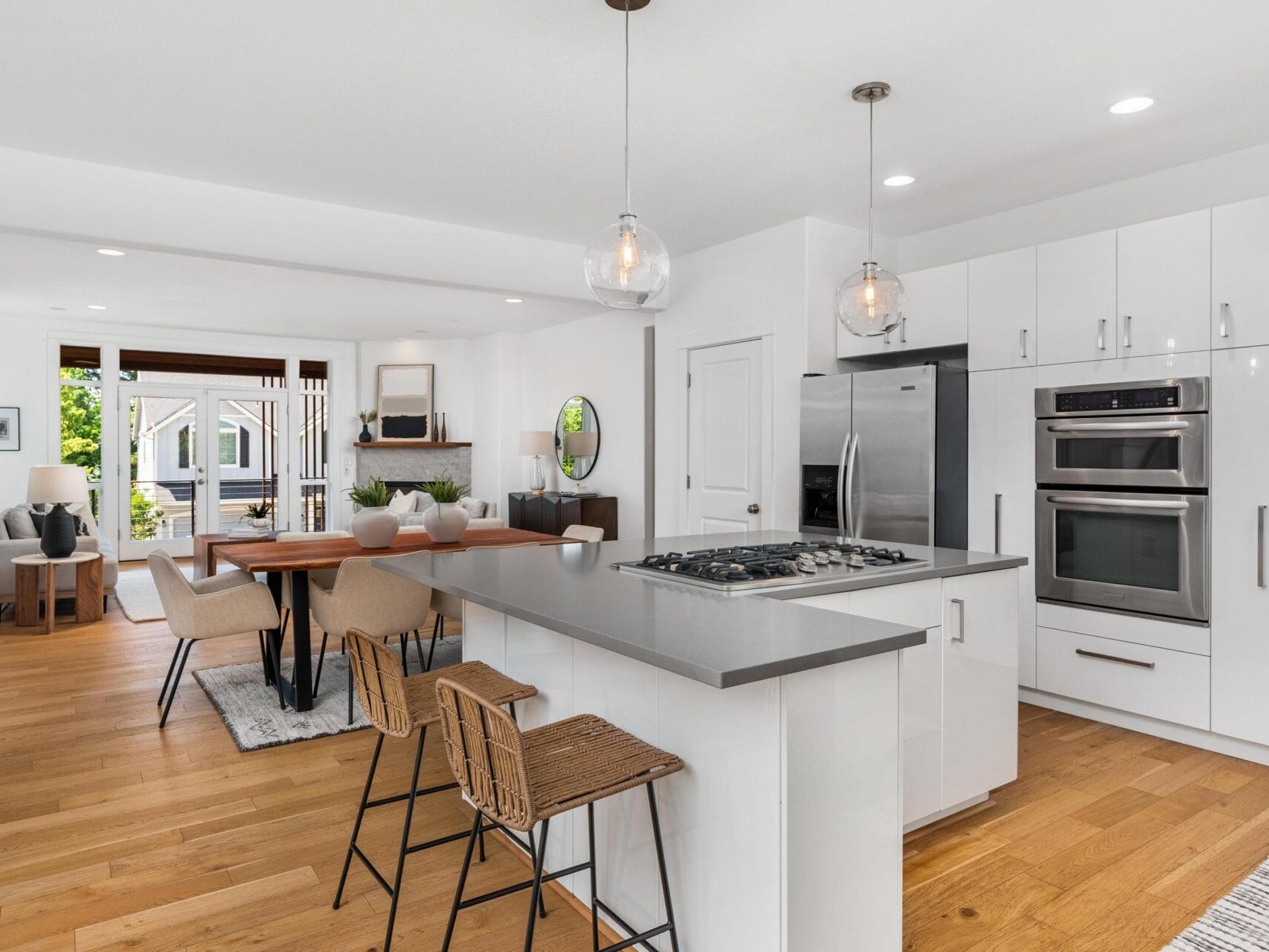 Modern open-concept kitchen and dining area with white cabinets, stainless steel appliances, island with bar stools, wooden dining table, and a cozy living space in the background with natural light.