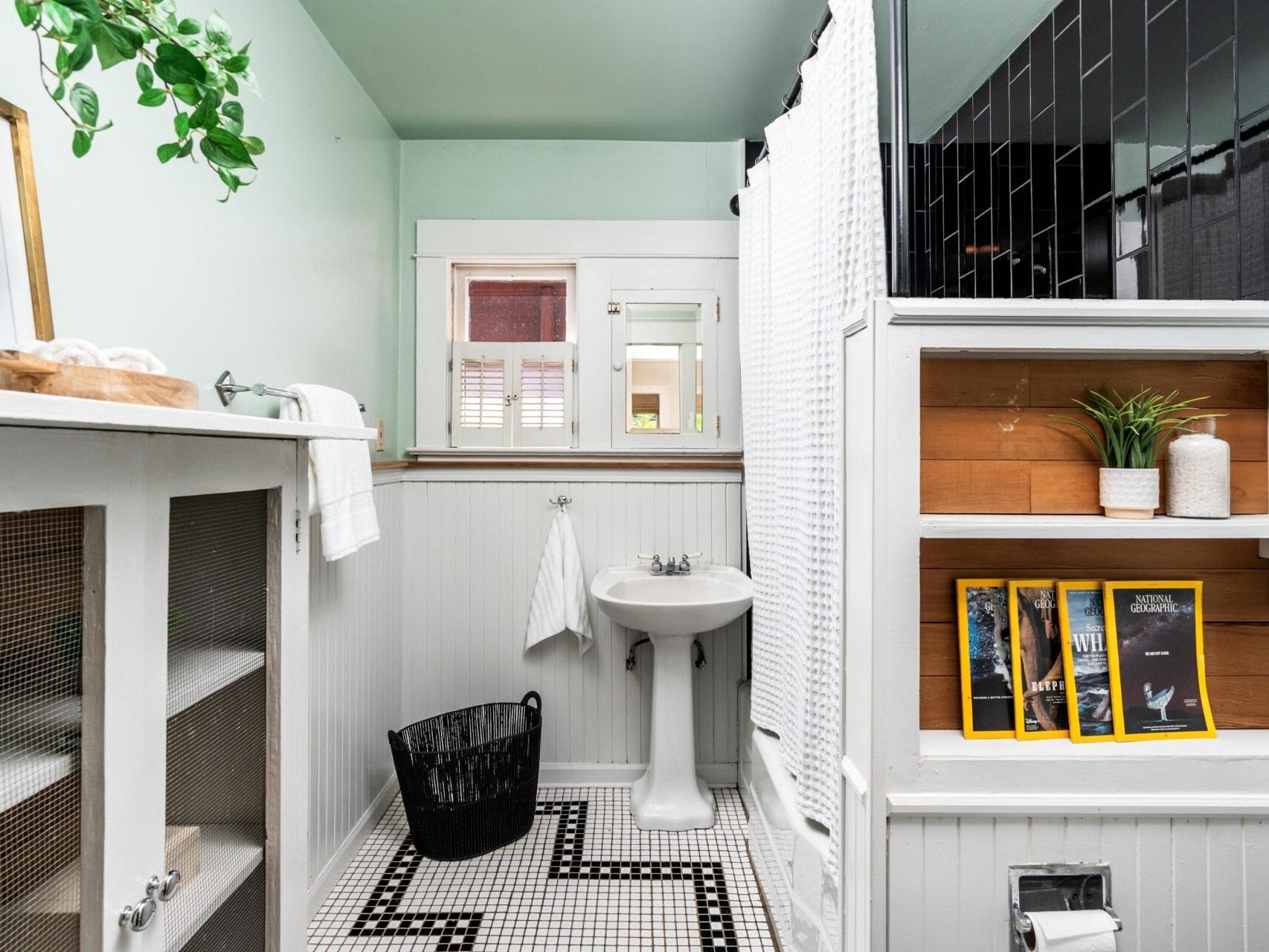 A bright, clean bathroom with a pedestal sink, white wainscoting, patterned black-and-white tile floor, a shower with white and black tiles, and neatly displayed magazines and decor on open shelves.