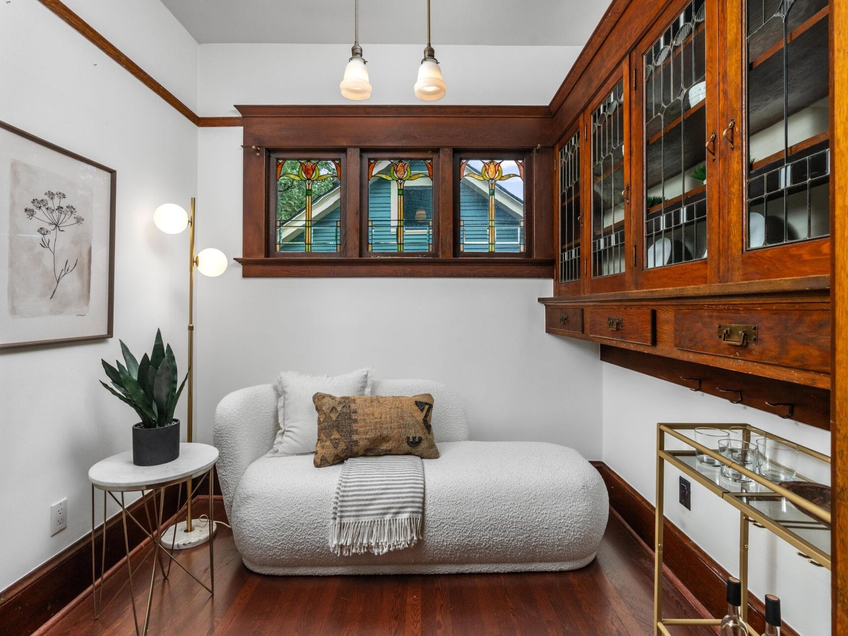 A cozy nook with a white textured loveseat, decorative pillows and blanket, a small round side table with a potted plant, stained glass windows, wooden built-in cabinets, and a gold bar cart on wood flooring.