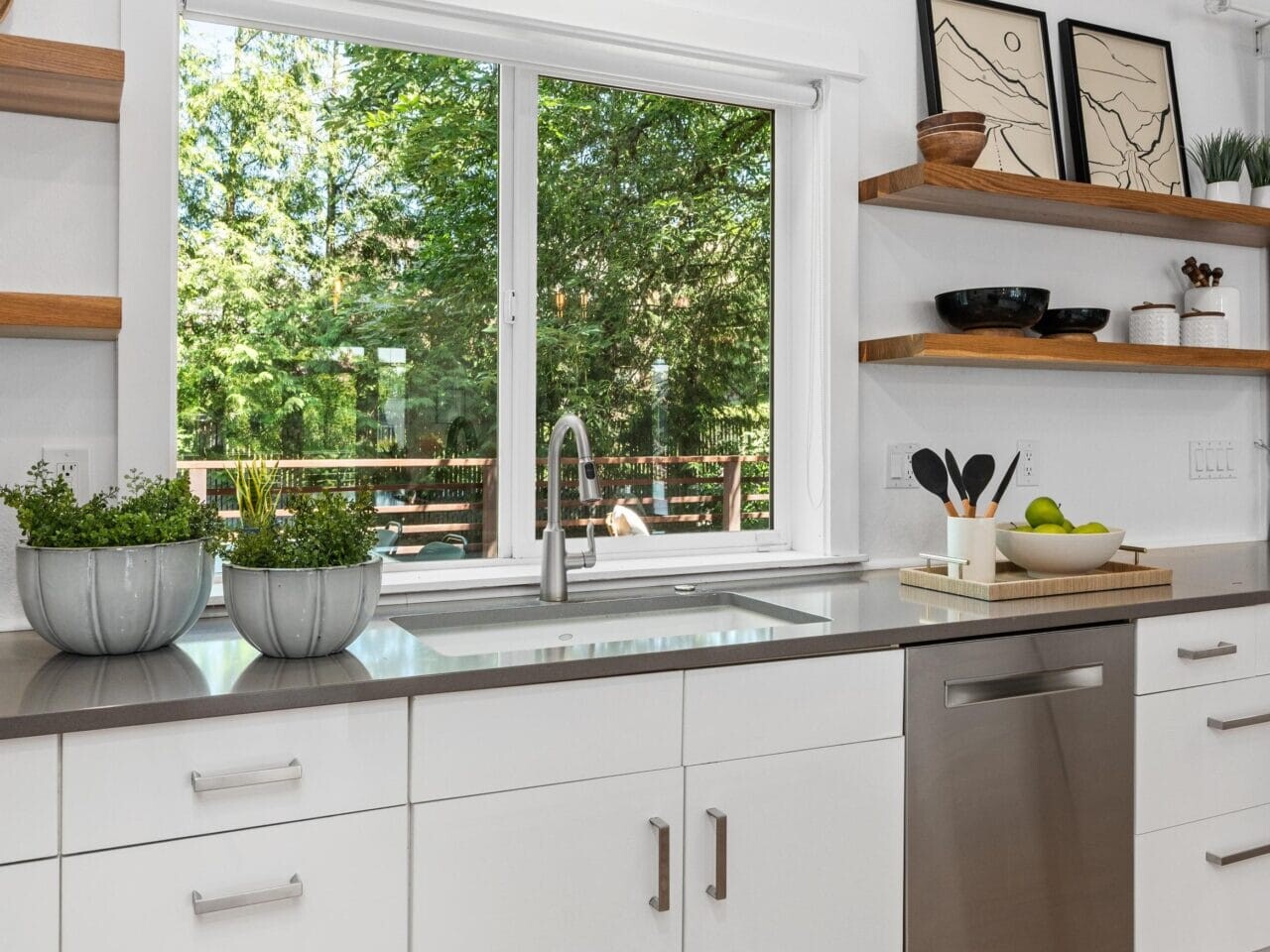 Bright modern kitchen with white cabinets, gray countertop, stainless steel dishwasher, potted herbs by the sink, floating wood shelves with dishes and decor, and a large window overlooking green trees.