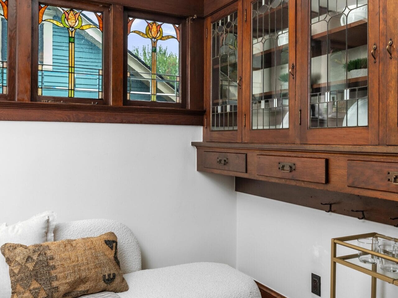 A cozy nook with a white textured sofa, a patterned pillow, and a striped throw. Stained glass windows let in light above built-in wooden cabinets. A small bar cart sits on the hardwood floor.