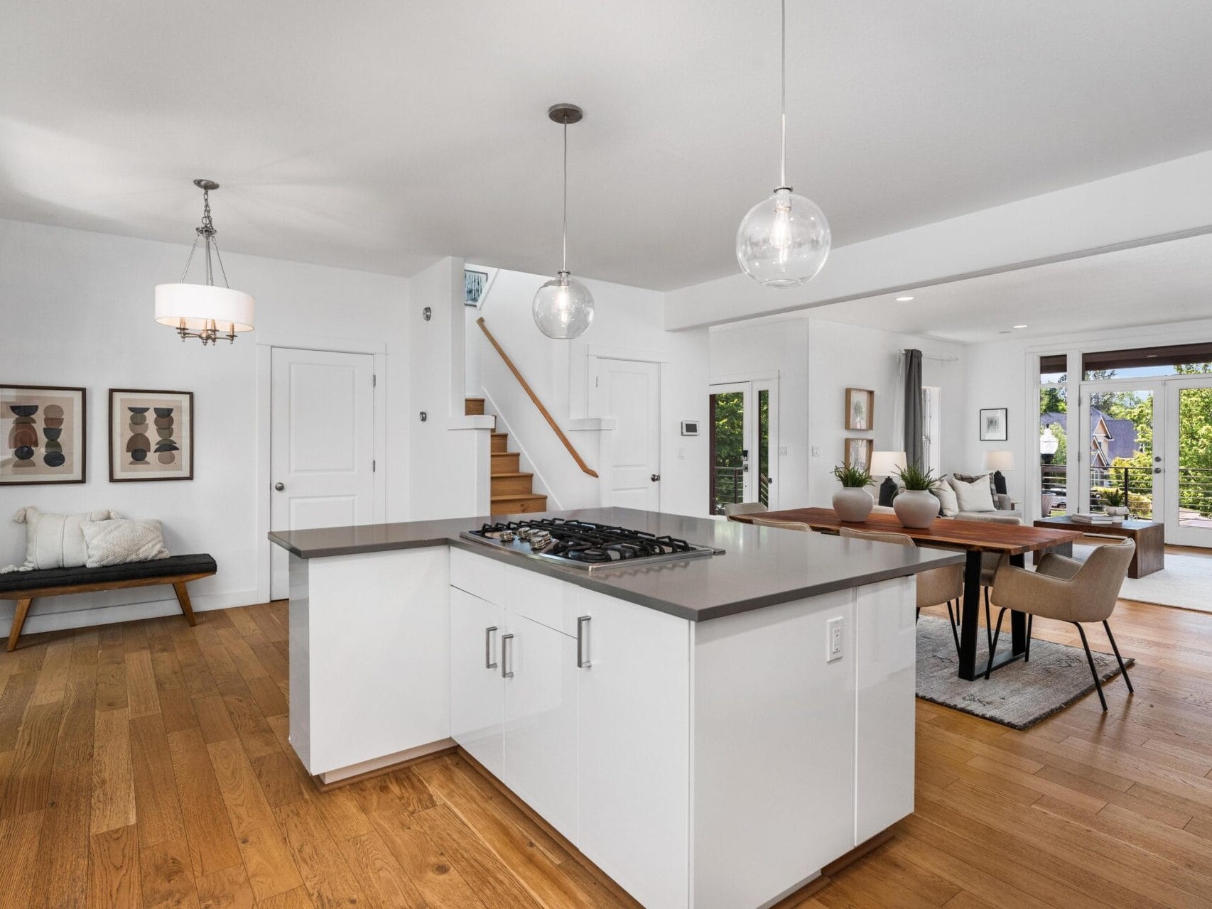 Modern open-concept kitchen and dining area with white cabinets, a central island with a cooktop, wooden floors, pendant lights, a dining table, and large windows letting in natural light.
