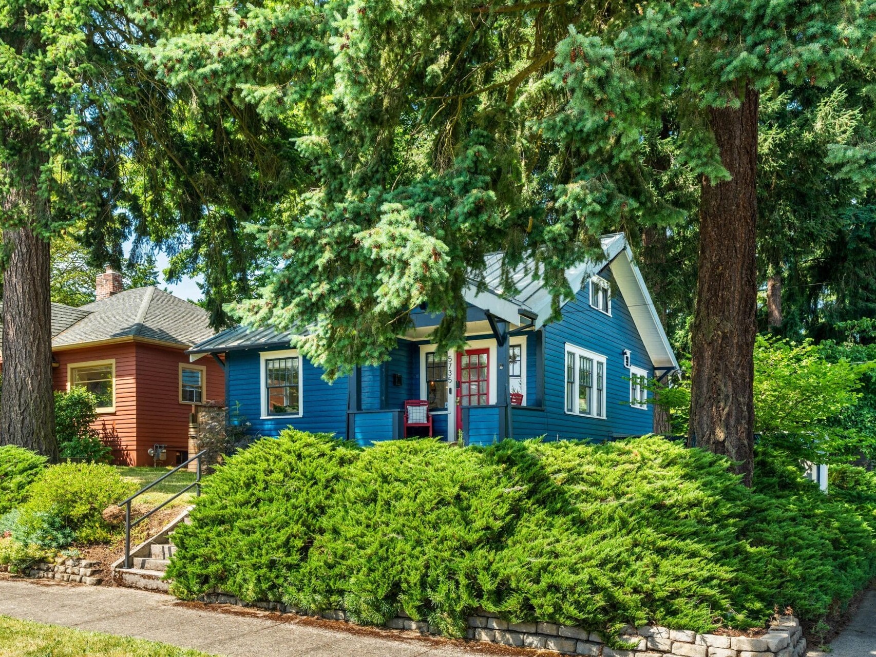 A blue house with a red door and white trim sits among tall trees and lush green bushes, next to an orange house, on a sunny residential street.