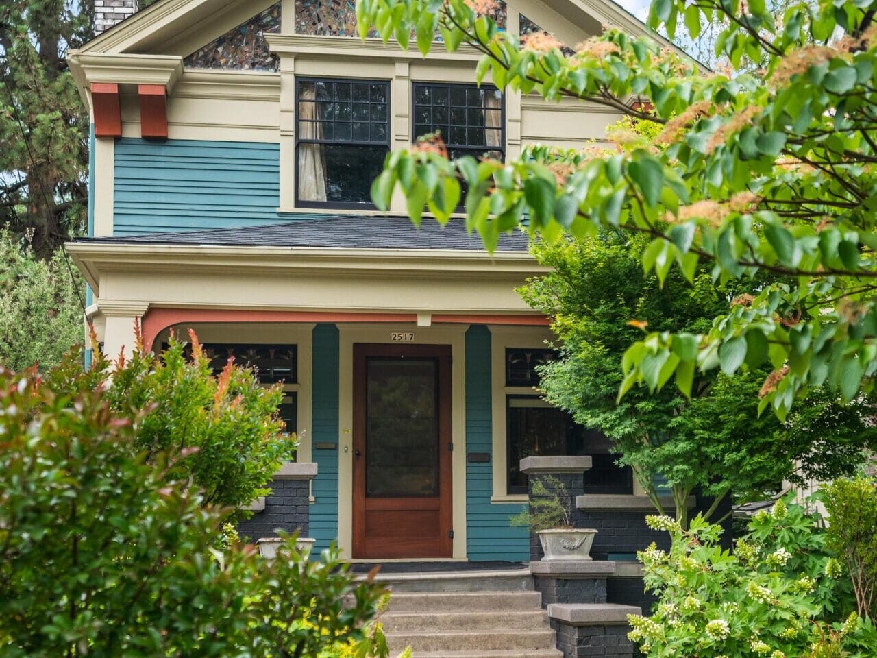A two-story house with blue siding, cream trim, and stained glass windows, partially obscured by lush green trees and shrubs, with concrete steps leading up to a wooden front door.