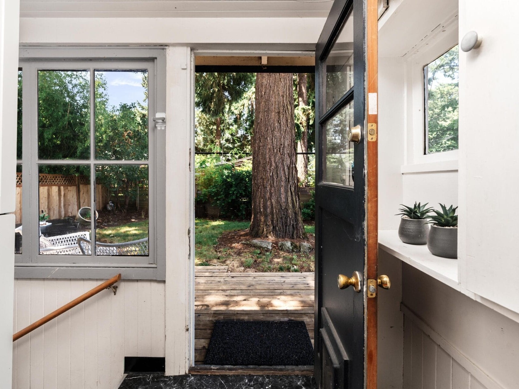A view from inside a house looking out through an open door onto a wooden deck and backyard with a large tree, greenery, and a fence; potted plants sit on a shelf by the door.
