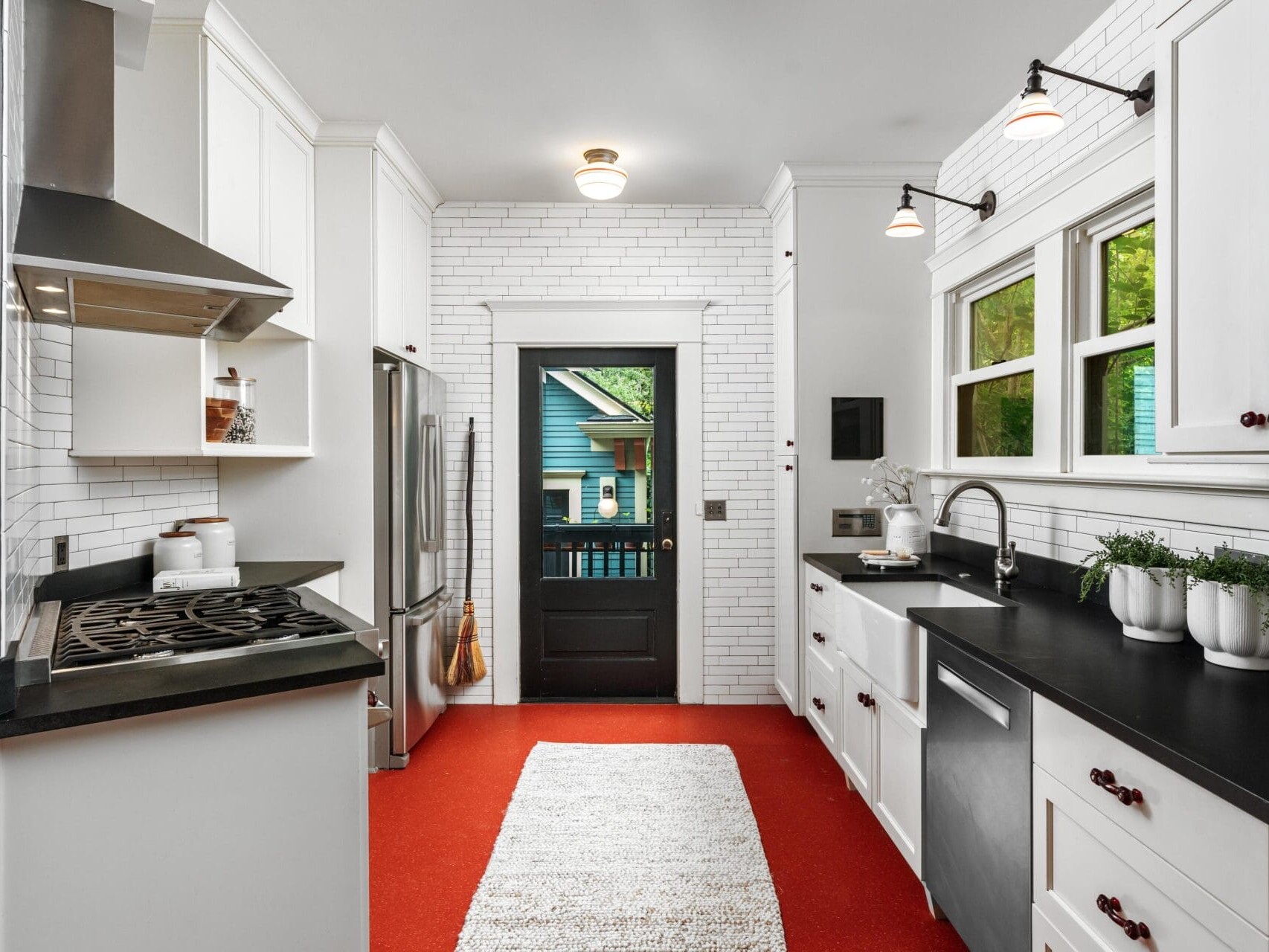 Modern kitchen with white cabinets, black countertops, red flooring, stainless steel appliances, and a white tile backsplash. There are windows over the sink and a door leading outside. White rug on the floor, potted plants by the sink.