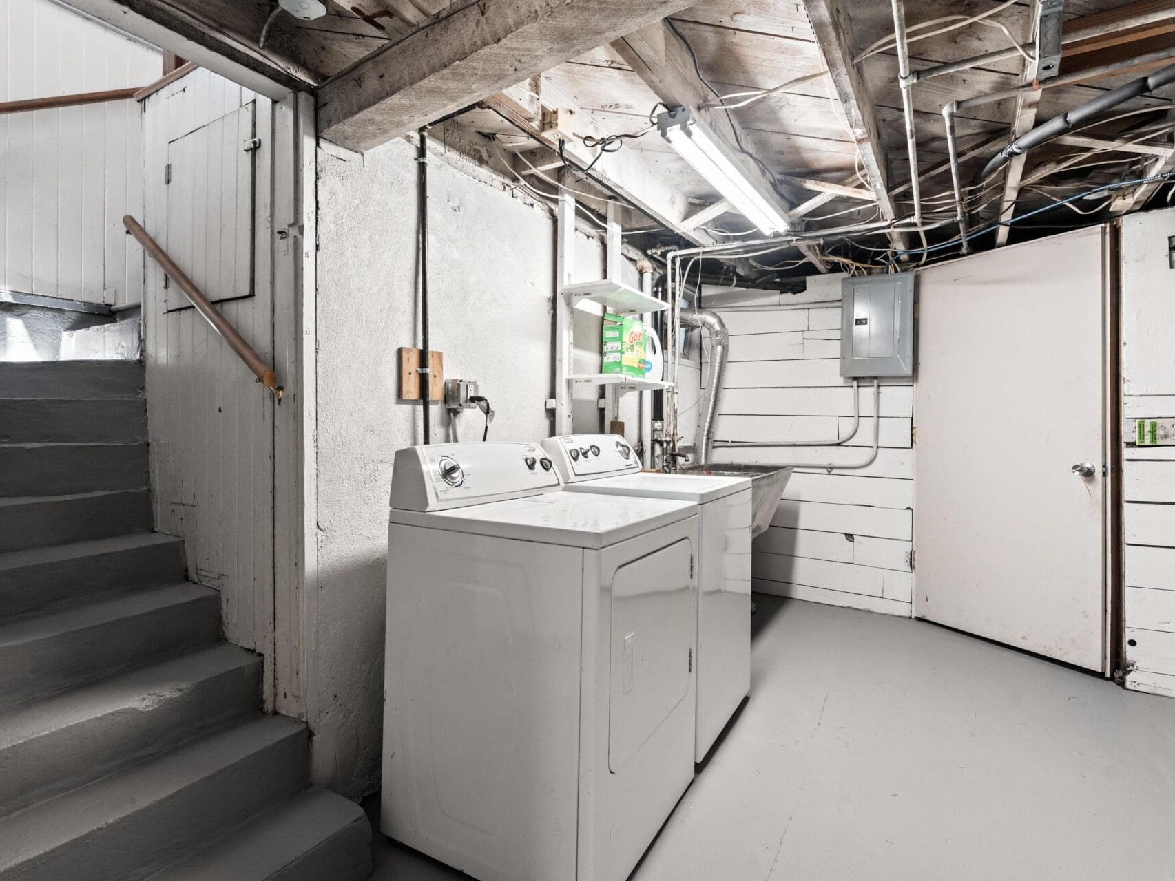 A basement laundry room with exposed beams and pipes, concrete floor, a white washer and dryer, shelves with detergent, a utility sink, electrical panel, and stairs leading upward. Walls are painted white.