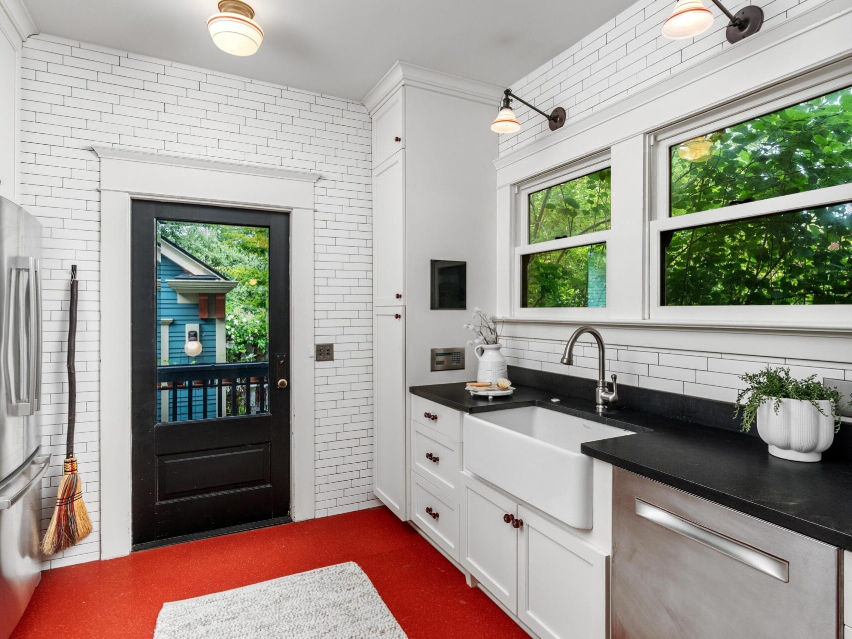 Bright kitchen with white cabinets, black countertops, farmhouse sink, stainless steel appliances, red floor, white subway tile walls, and large windows showing lush green trees outside. A door leads to a porch.