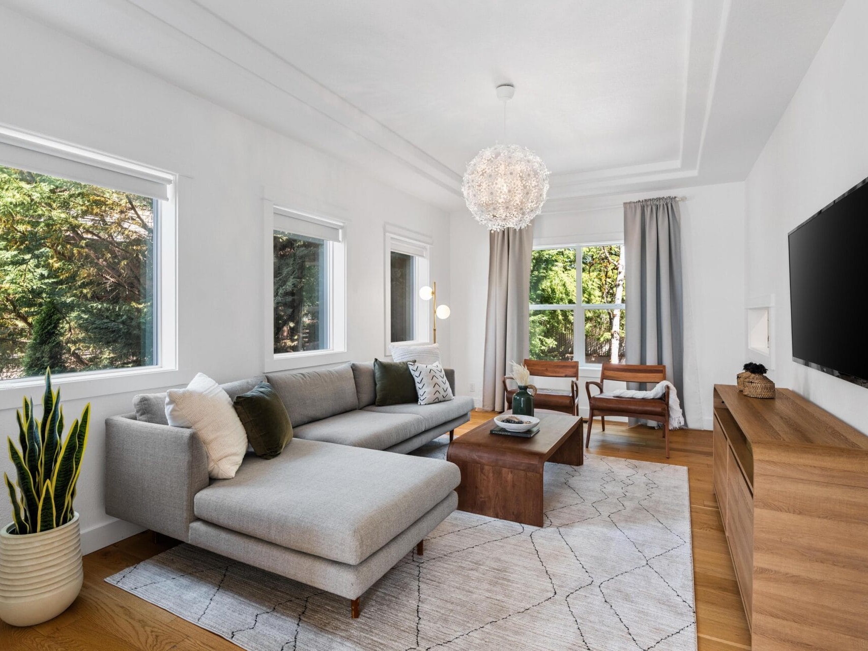 A modern living room with a grey sectional sofa, wooden coffee and media tables, TV, large windows, a geometric rug, green plant, and a statement chandelier. Natural light fills the space with neutral decor accents.