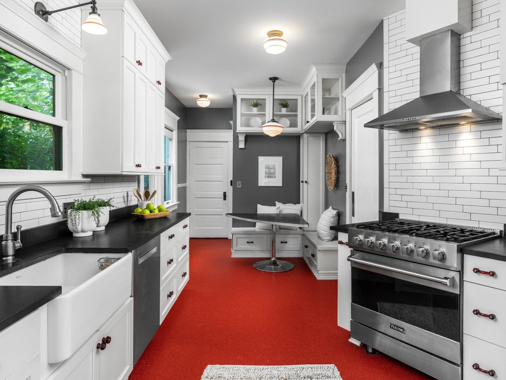 Modern kitchen with white cabinets, black countertops, stainless steel appliances, farmhouse sink, and a bright red floor. There is a cozy breakfast nook with benches and large windows providing natural light.