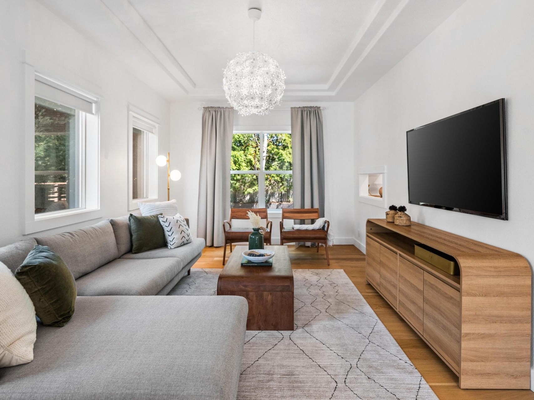 Modern living room with a gray sectional sofa, wooden coffee table, flat-screen TV on a wooden console, and a dining table near large windows with gray curtains. White walls and a contemporary chandelier complete the space.