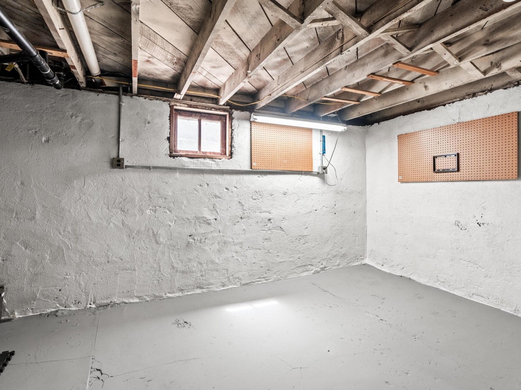 A bare basement room with white painted stone walls and floor, exposed wooden ceiling beams, small windows, and pegboards mounted on two walls. Light streams in from the small window.