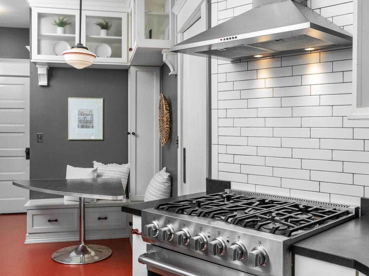 Modern kitchen with a stainless steel gas stove and hood, white subway tile backsplash, black countertops, red flooring, and a built-in breakfast nook with gray and white decor.