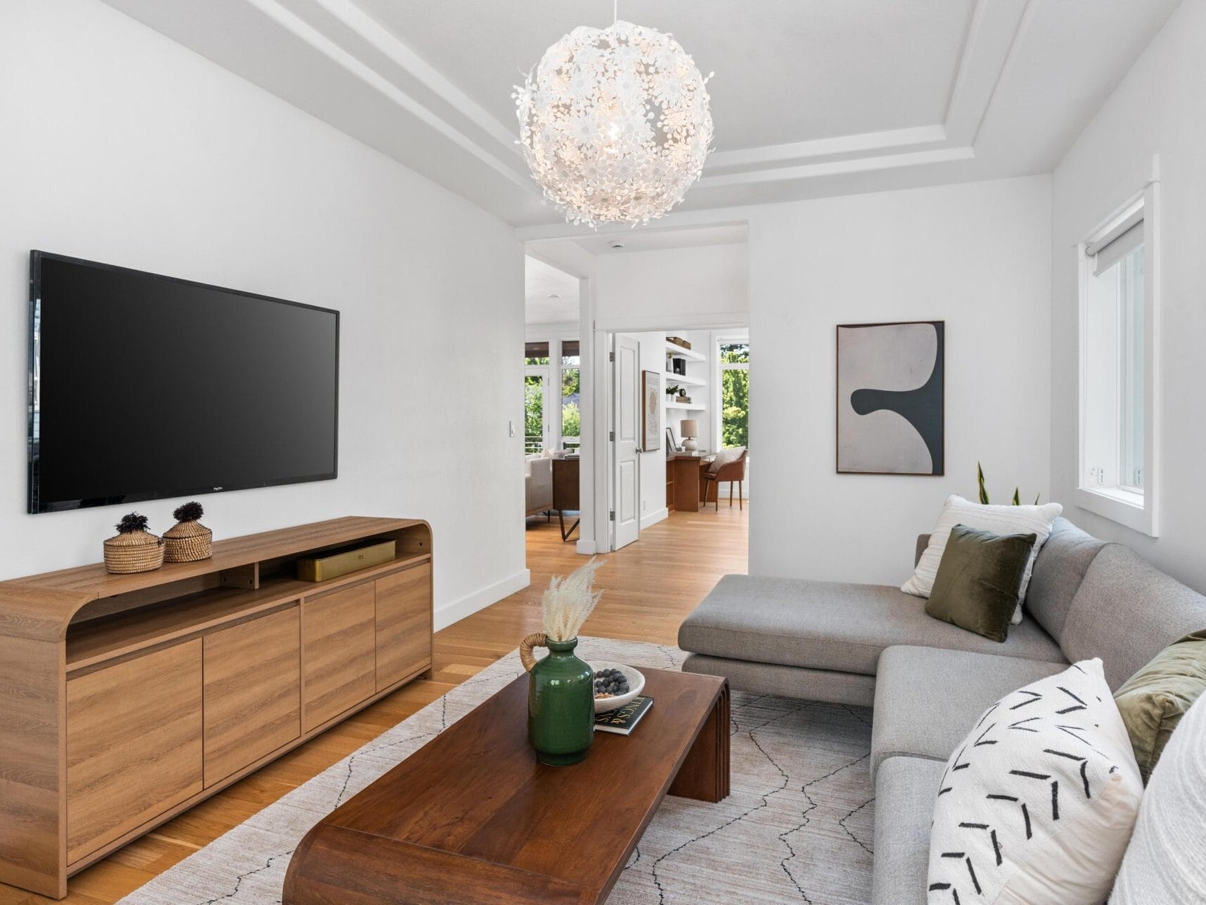 Modern living room with a gray sectional sofa, abstract wall art, wooden coffee table, and TV mounted on a wooden media console. White walls, wood flooring, large windows, and a decorative chandelier complete the space.