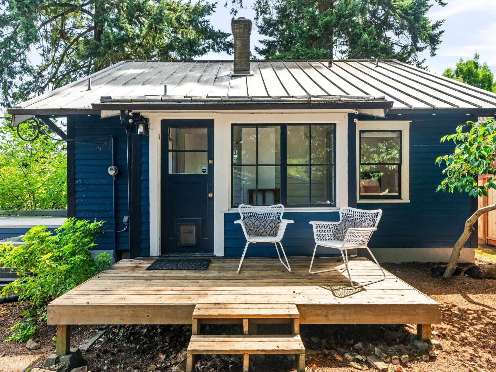 Small blue house with a metal roof, large window, and a front porch featuring two white chairs. Surrounded by trees and greenery, the porch has wooden steps leading to a simple garden area.