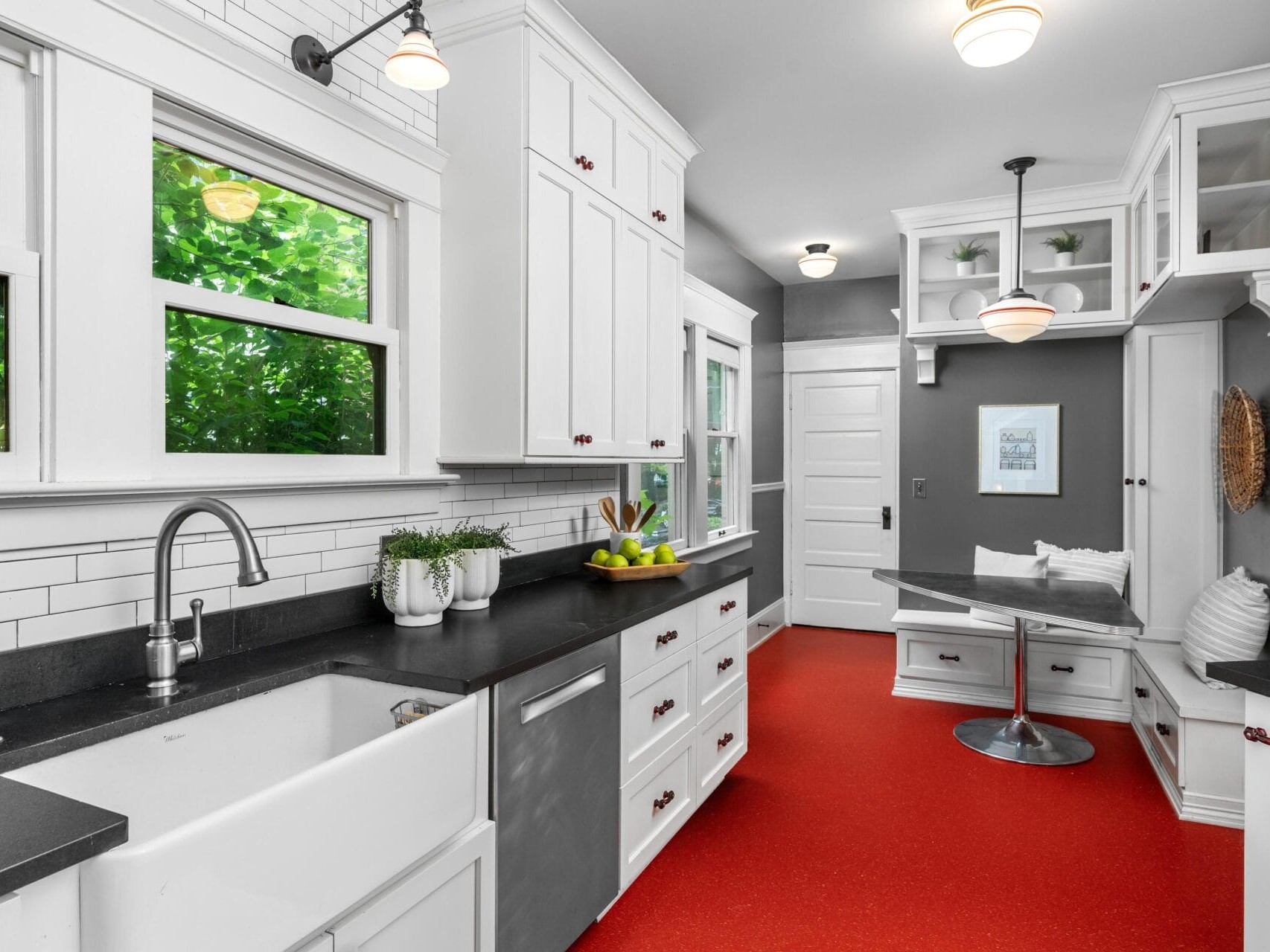 Bright kitchen with white cabinets, black countertops, farmhouse sink, stainless appliances, red floor, subway tile backsplash, large windows, and a cozy built-in nook with a table and bench. Greenery visible outside the windows.
