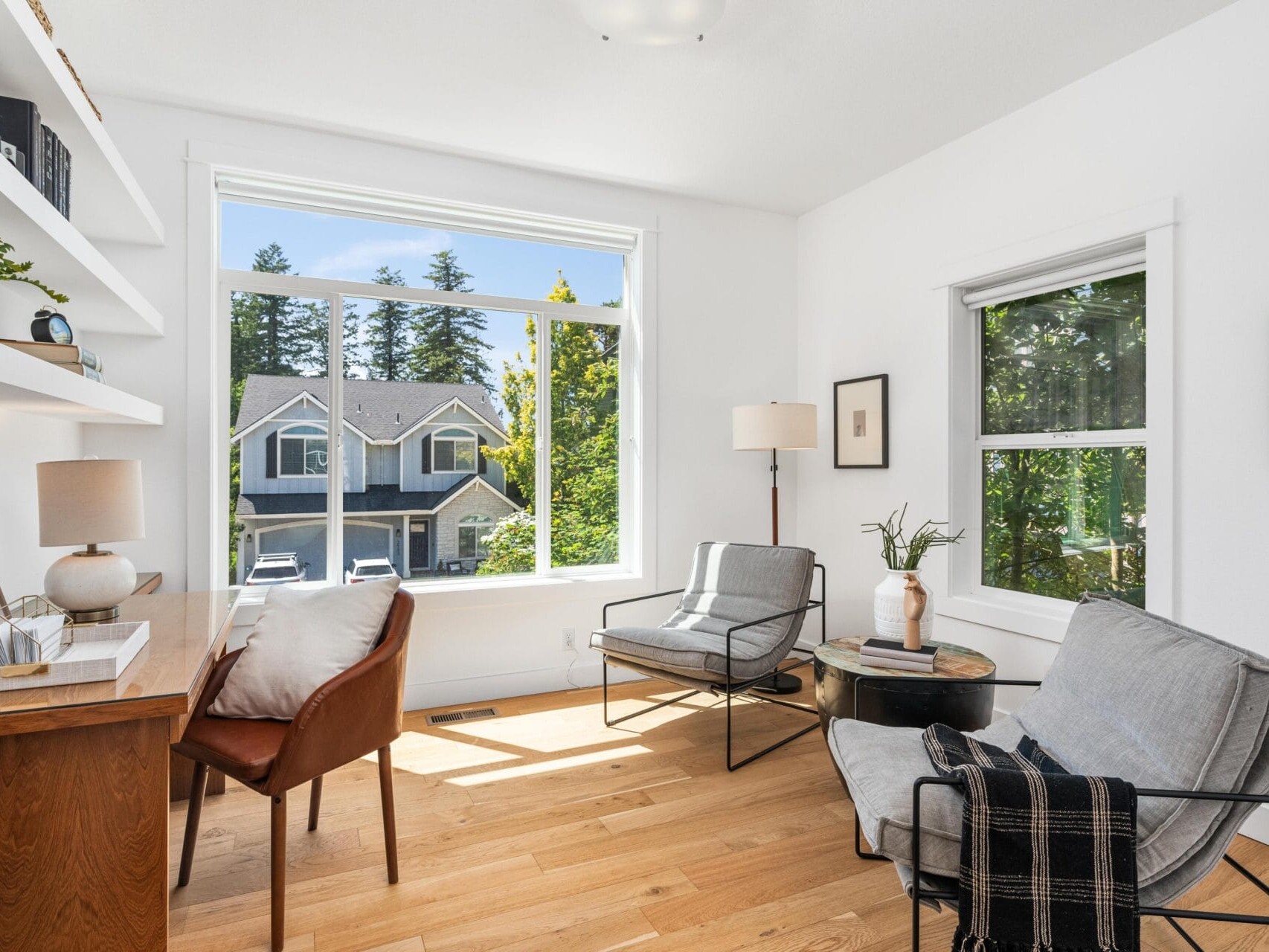 Bright home office with a wooden desk, brown leather chair, two modern armchairs, and large windows showing a view of trees and houses outside. Shelves and plants add decor to the airy, sunlit room with light wood floors.