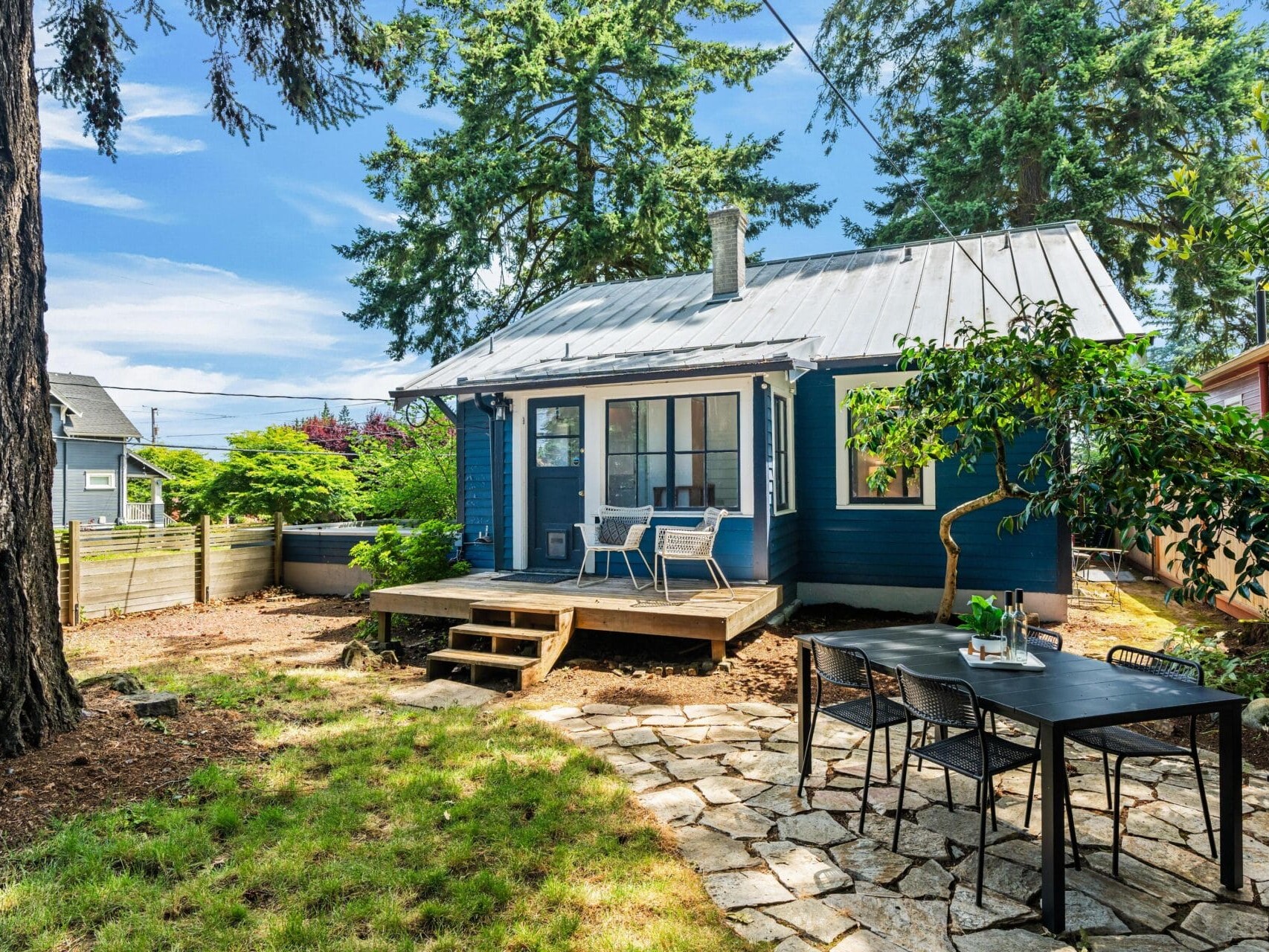 A small blue house with a metal roof and a wooden porch sits in a shady backyard with patio furniture, a stone dining area, and surrounded by tall trees and a wooden fence.