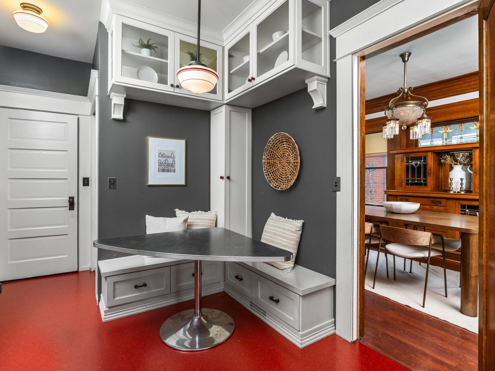A cozy breakfast nook with a black tabletop, white benches, and gray walls sits next to a dining room with wood furniture, separated by a sliding door. Red flooring contrasts with white trim and cabinetry.