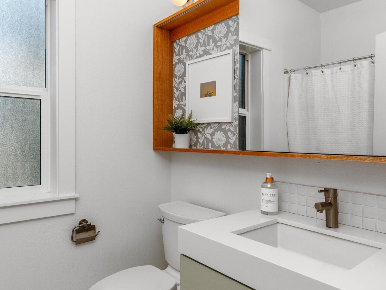 Modern bathroom with white walls, a window, toilet, and a sink with a green vanity. A wooden-framed mirror with shelves is above the sink, and a shower with a white curtain is in the background.