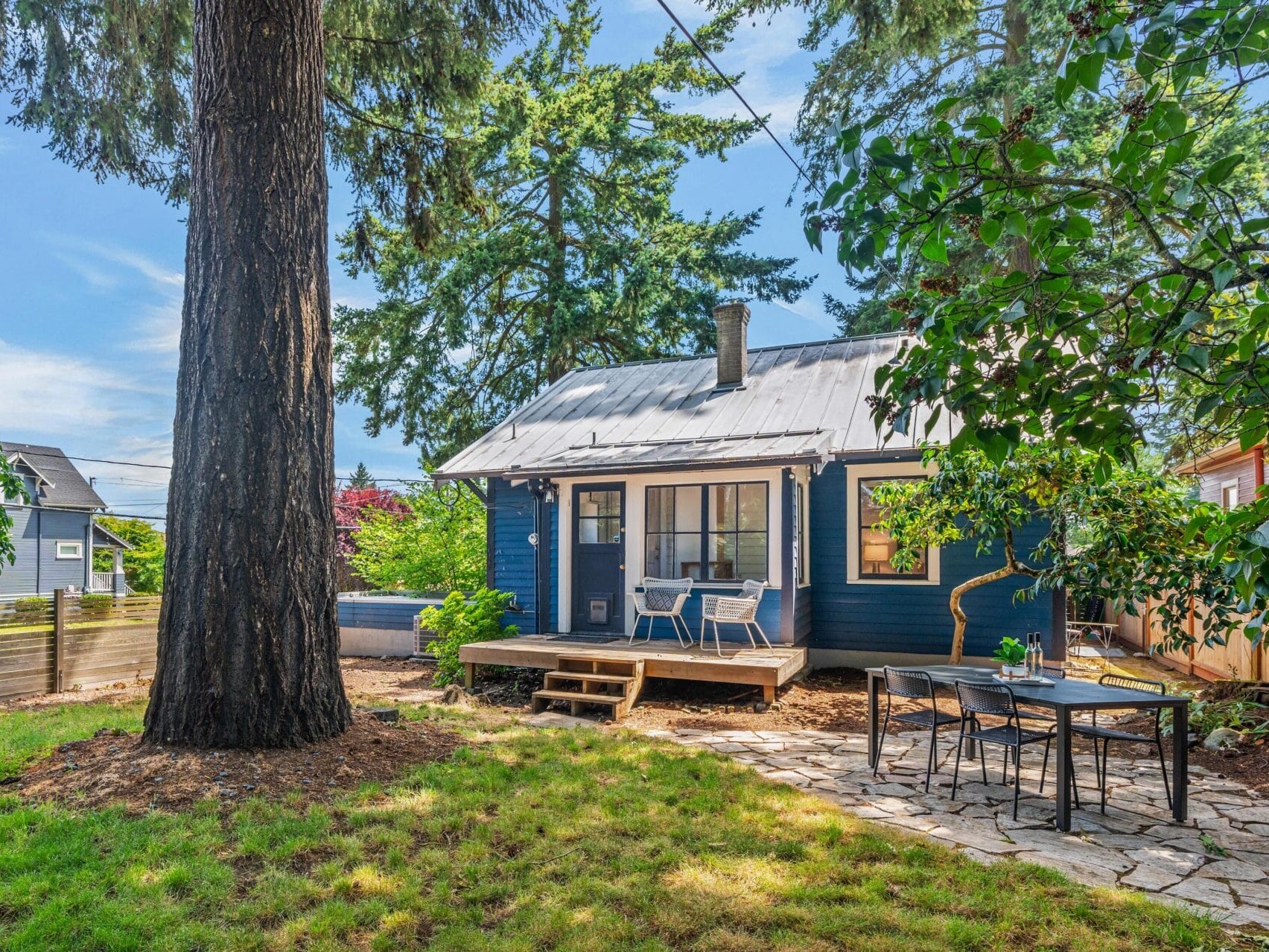 A cozy blue cottage with a metal roof sits in a backyard surrounded by trees and greenery. There’s a wooden deck with chairs, a stone patio with an outdoor dining table, and sunlight filtering through the branches.