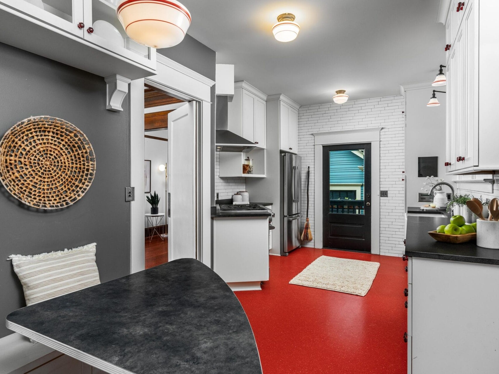 Modern kitchen with red flooring, white cabinets, and dark countertops. A woven wall decoration and a black table with a striped cushion are in the foreground. Door at the back opens to an exterior view. Appliances and decor are visible.