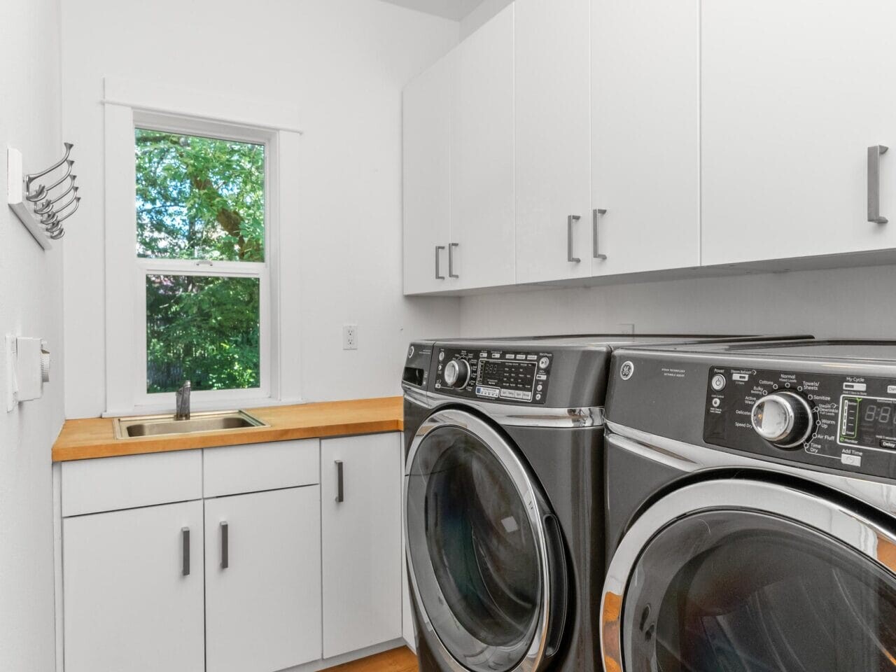 A modern laundry room with white cabinets, a wooden countertop, a sink, a window with a green view, and a black front-loading washer and dryer on hardwood flooring.