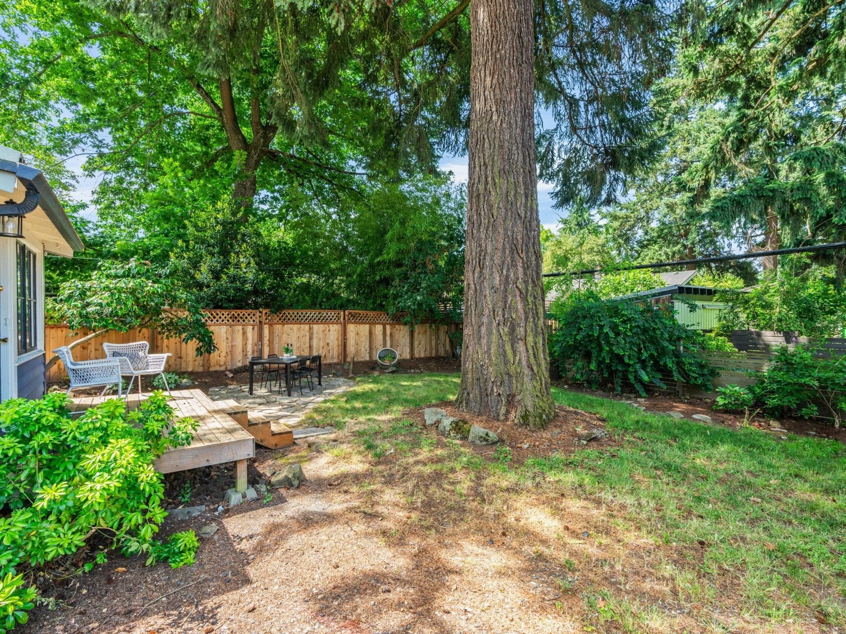 A sunny backyard with a large tree, wooden deck with patio chairs, a fire pit with seating, wooden fence, and lush greenery surrounding the area.
