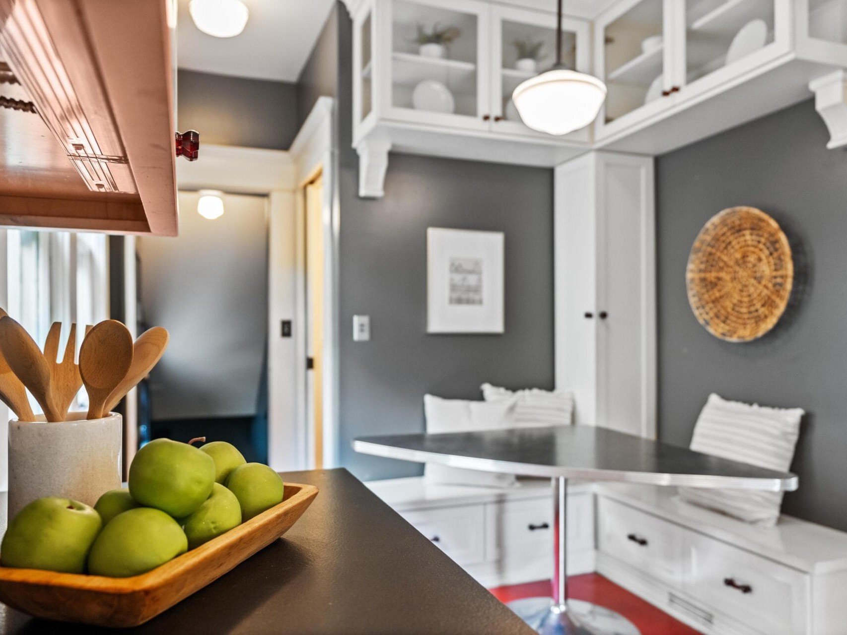 A modern kitchen nook with gray walls, built-in white benches and storage, a metal table, wall art, and a bowl of green apples beside wooden utensils on a countertop in the foreground.