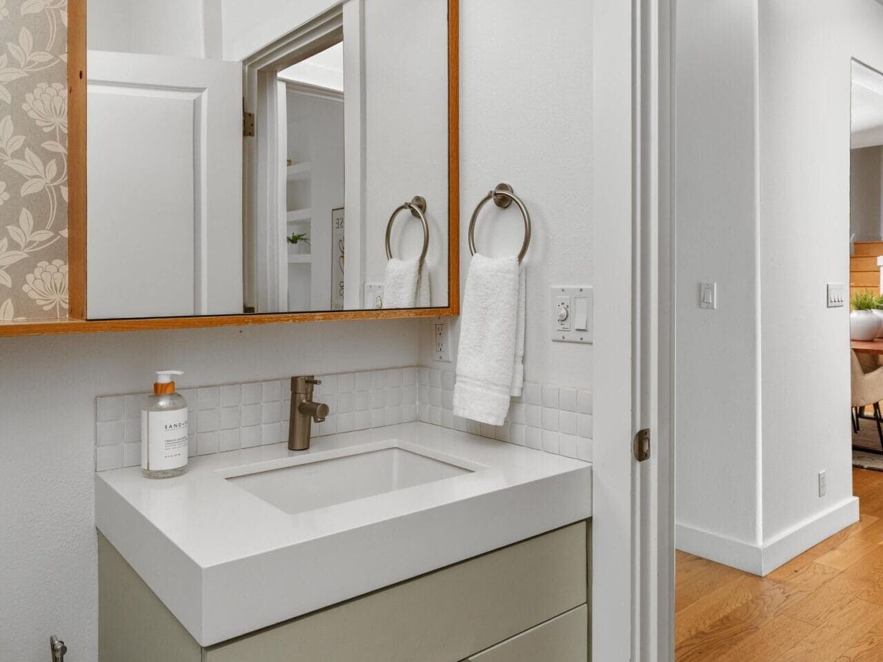 Modern bathroom with a light green vanity, white sink, wall-mounted faucet, hand towel on a ring, soap dispenser, mirrored cabinet, and wooden floors in the adjoining hallway.
