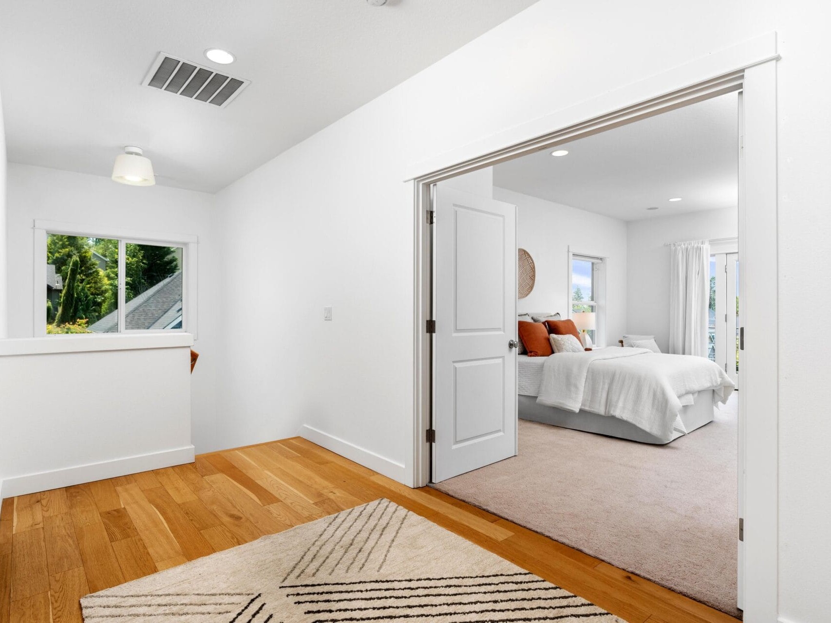 A bright hallway with wooden floors leads to a bedroom with carpet, a white bedspread, orange cushions, and large windows letting in natural light. Double doors separate the hallway from the bedroom.