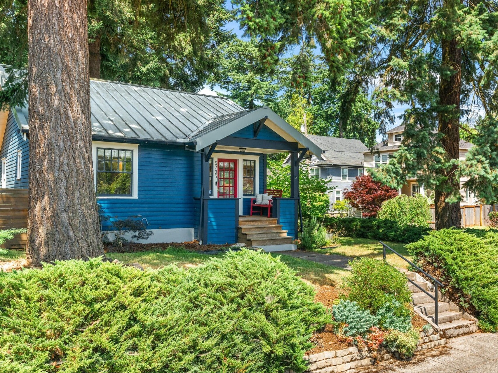 A small blue house with a metal roof and red front door, surrounded by tall trees and green shrubs, with a front porch and steps leading to the entrance on a sunny day.