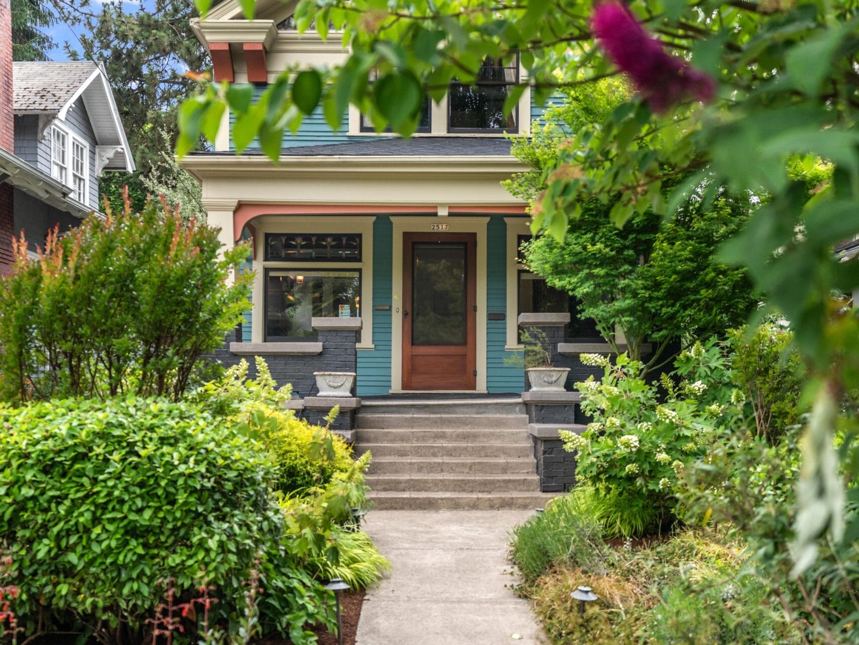 Front view of a charming house with green and blue trim, large windows, and a wooden front door. The entry is framed by lush greenery and bushes, with steps leading up from a concrete path.