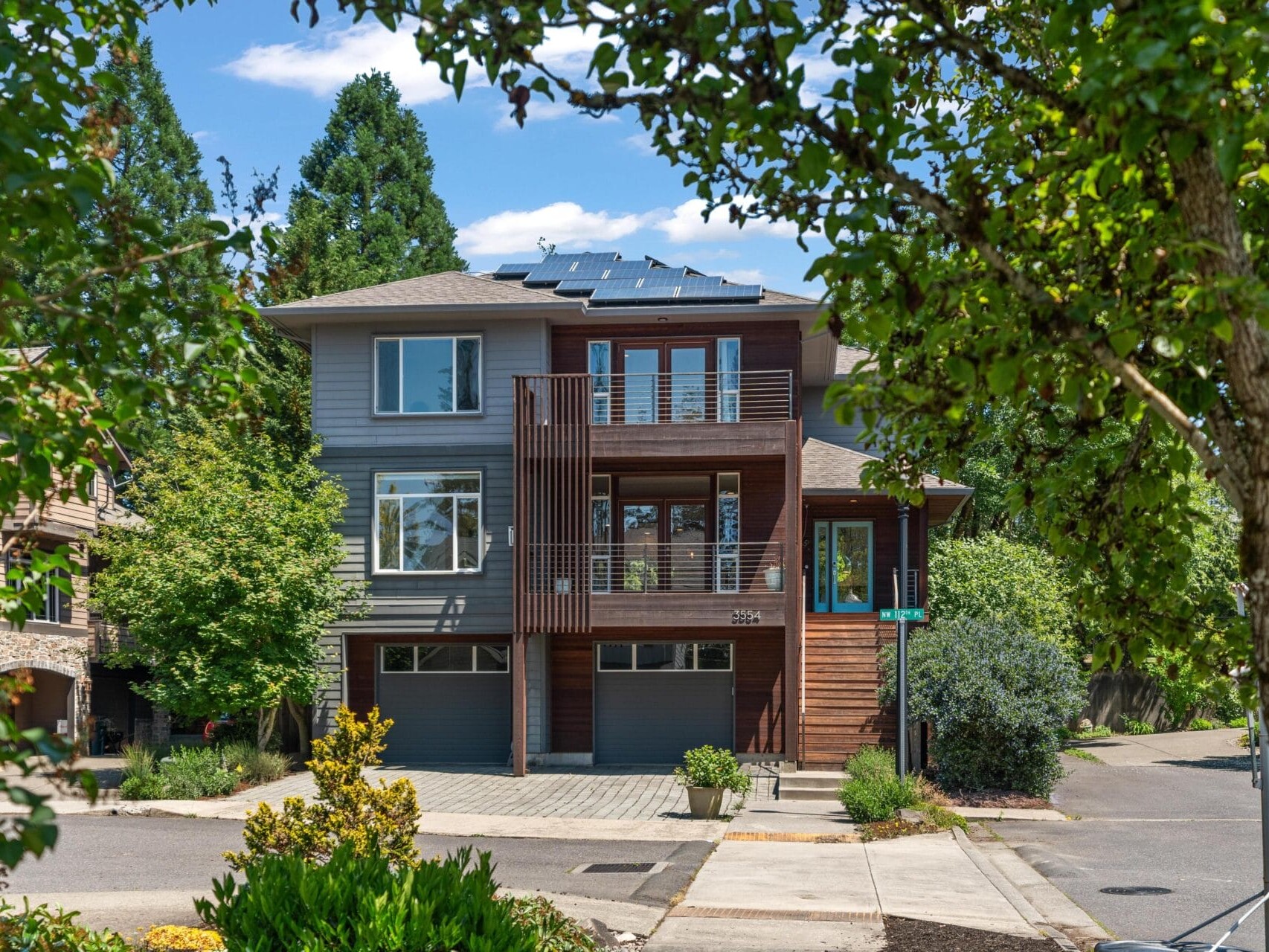 Modern three-story house with gray and wood exterior, large windows, and solar panels on the roof, surrounded by trees and greenery on a sunny day at a quiet street corner.