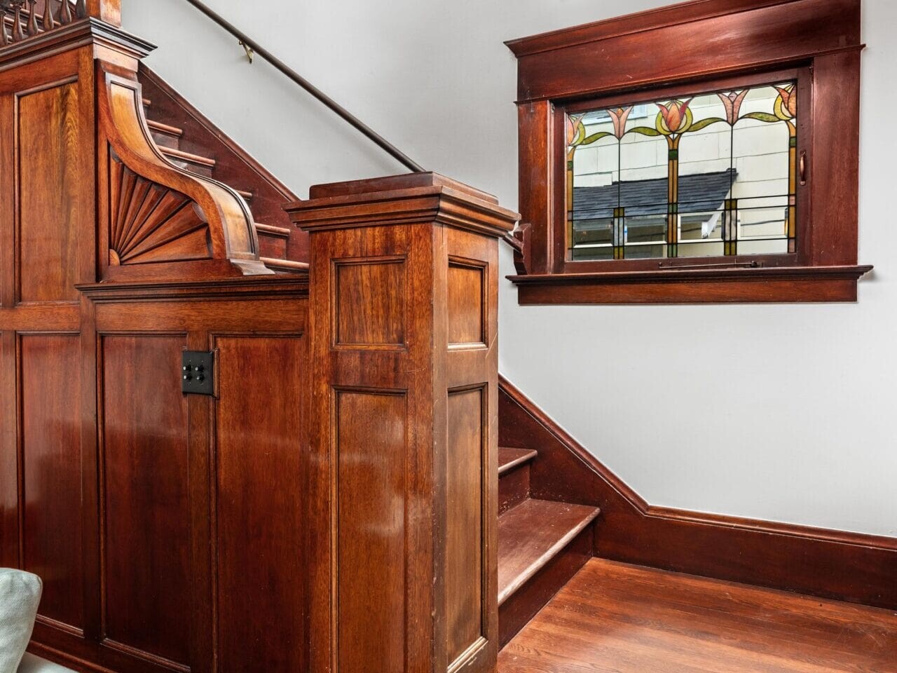Wooden staircase with ornate paneling and banister, featuring a stained glass window on the landing. Rich, dark wood tones contrast with white walls, creating a classic, elegant look.