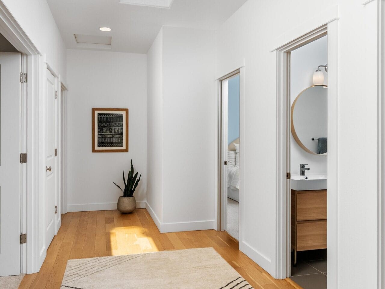 A bright hallway with light wood flooring, white walls, a patterned beige rug, and three open doors leading to other rooms. A potted plant and framed art are at the end of the hall under a skylight.